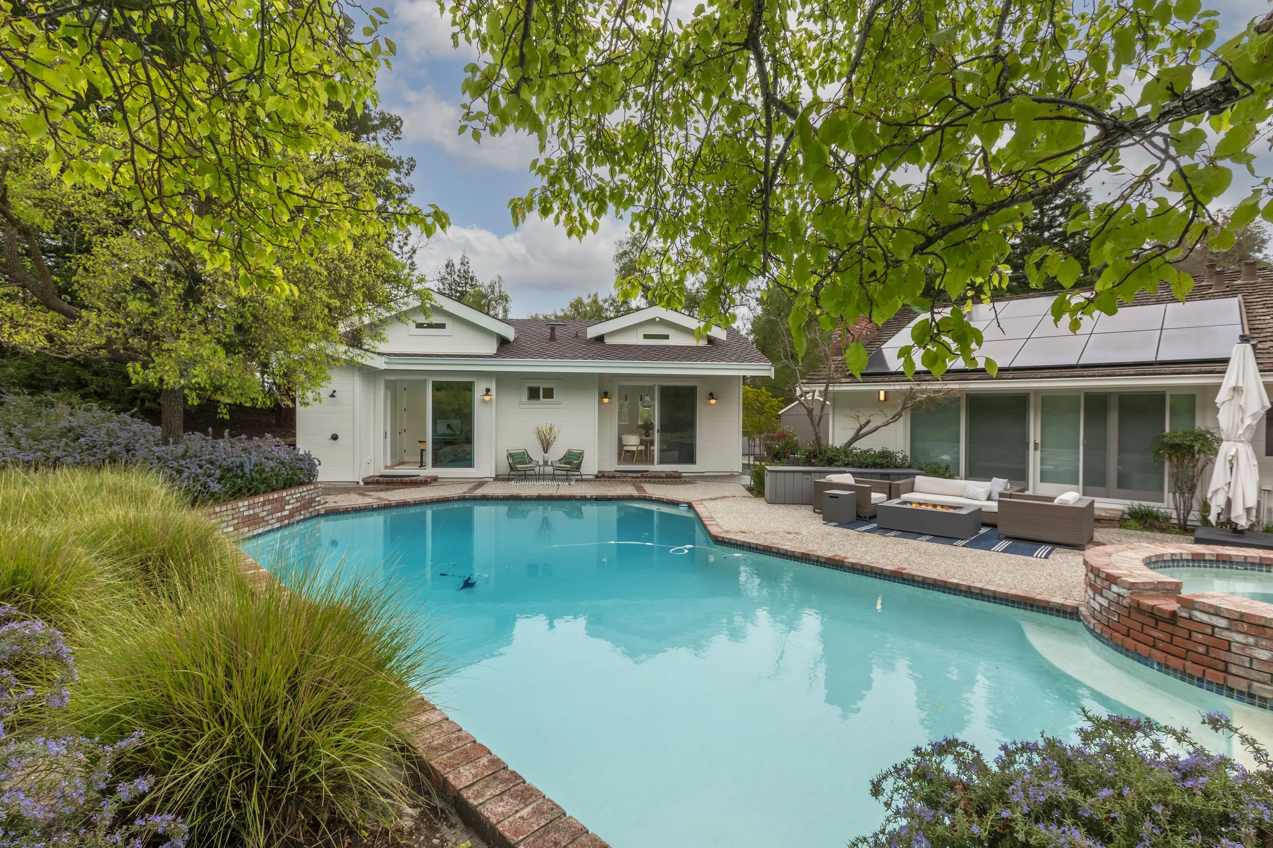 A backyard with a swimming pool surrounded by plants and outdoor furniture, with a white house featuring solar panels on the roof in the background, and trees with green leaves overhead.