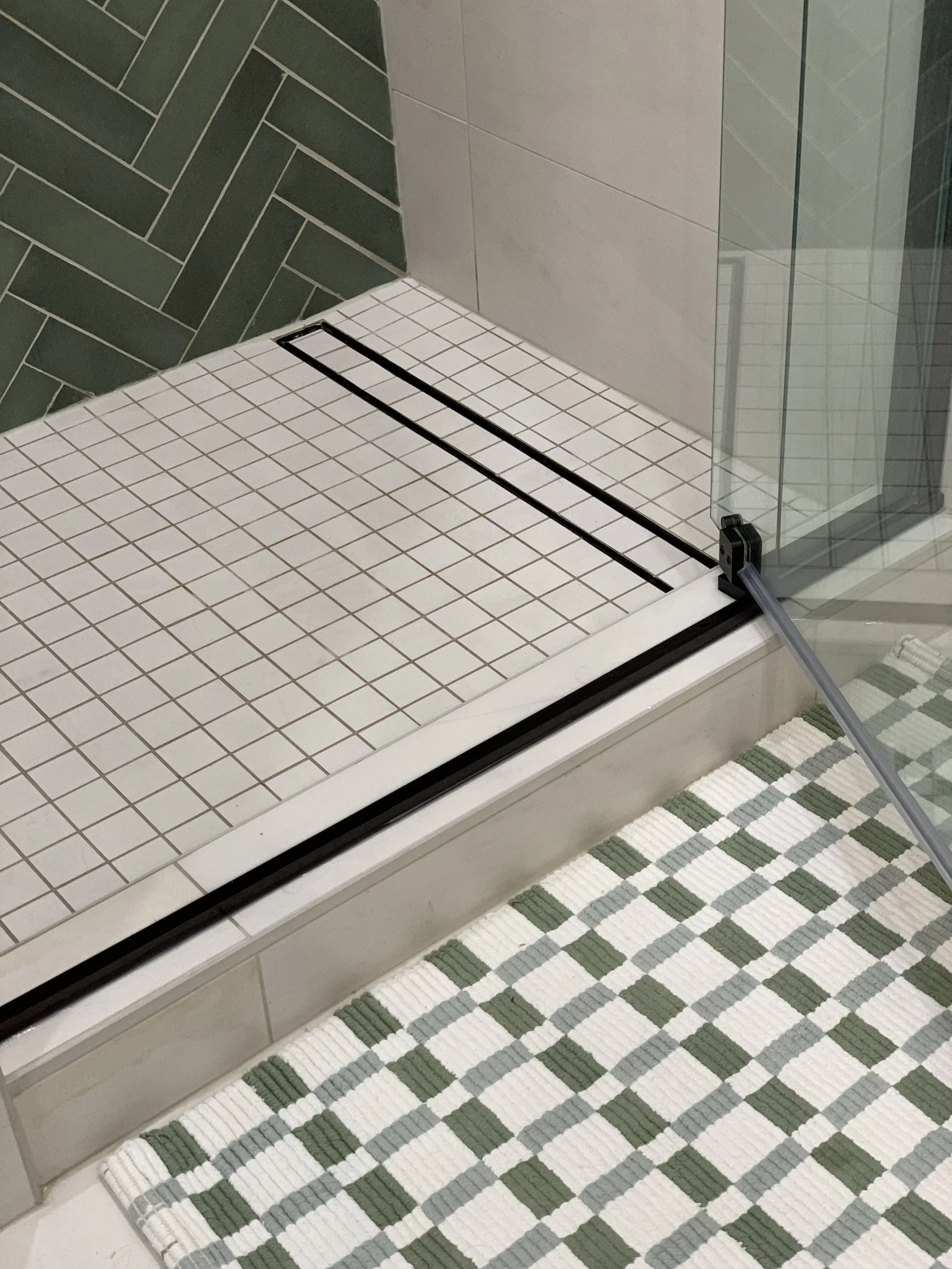 The image shows a close-up view of a bathroom shower area with a glass door. The floor is tiled with small white tiles. The shower drain is long and rectangular, bordered with black. The wall inside the shower has white tiles, and the adjacent wall o
