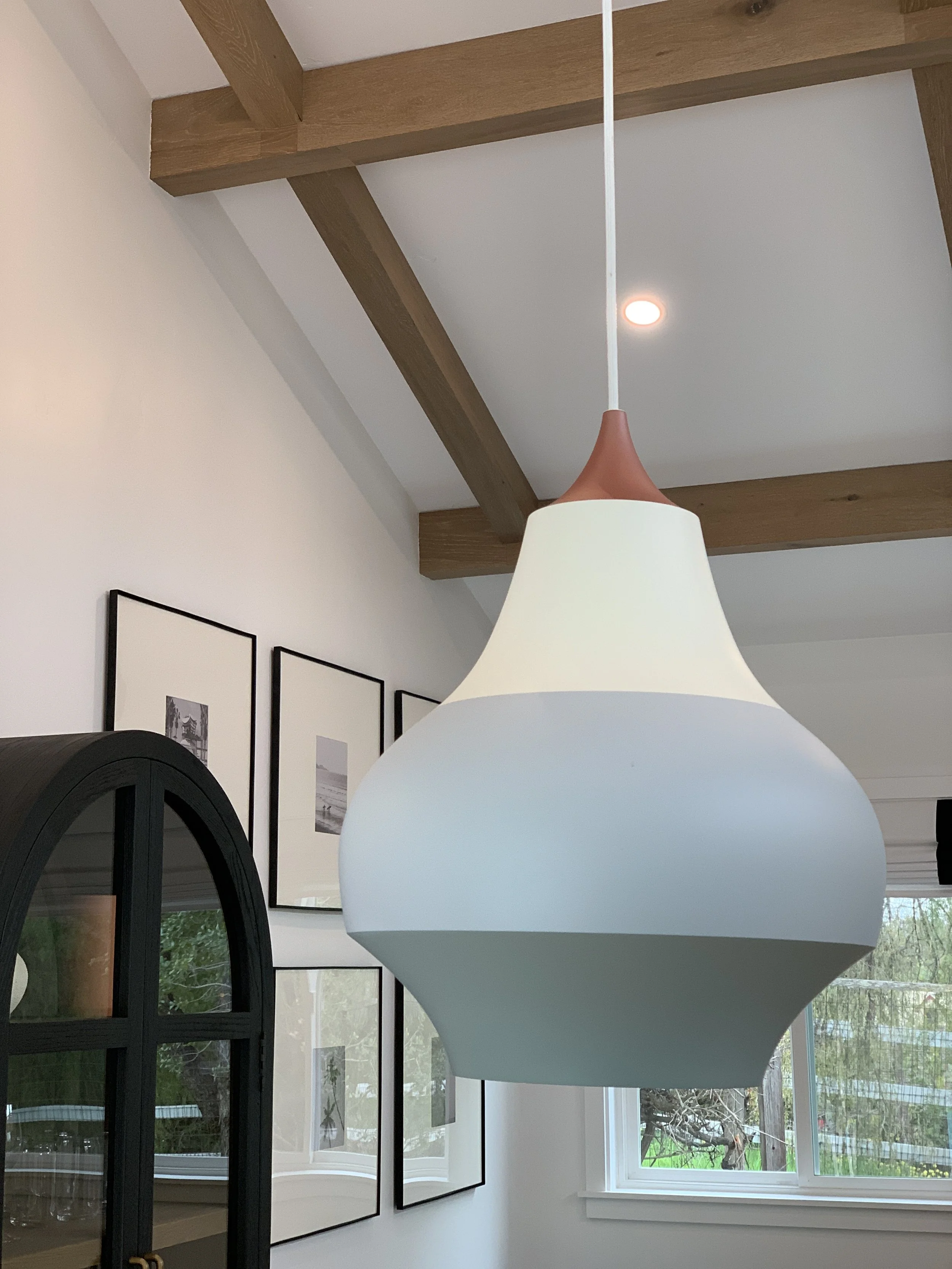 A modern living room with a white pendant light fixture hanging from the ceiling, wooden beams, and large windows showing greenery outside. Wall art with black frames and pictures are also visible.