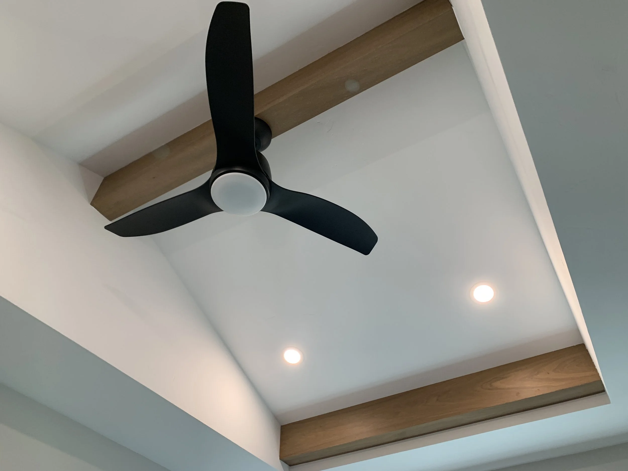 Ceiling fan with three black blades and white center, installed on a ceiling with wooden beams and recessed lighting.