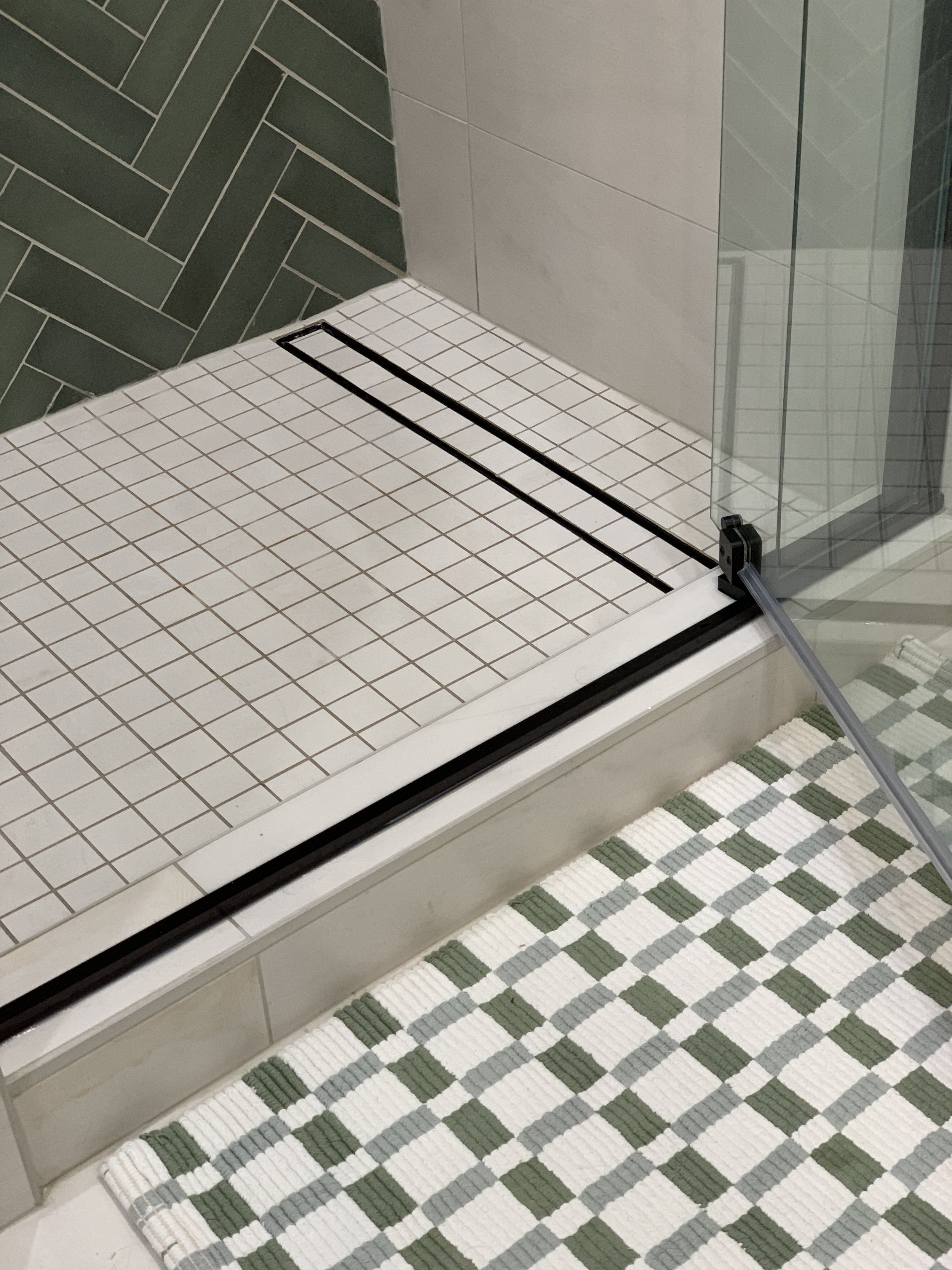 A bathroom shower area with a white tiled floor and a glass door. The wall tiles are a combination of green herringbone pattern and plain white tiles. There is a striped rug on the floor outside the shower.