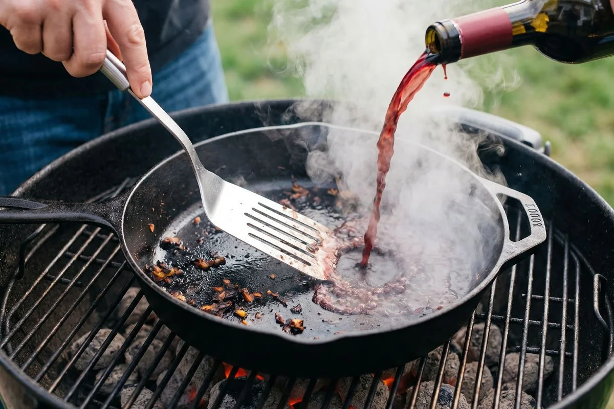 Deglazing a cast iron pan with red wine on a charcoal grill.