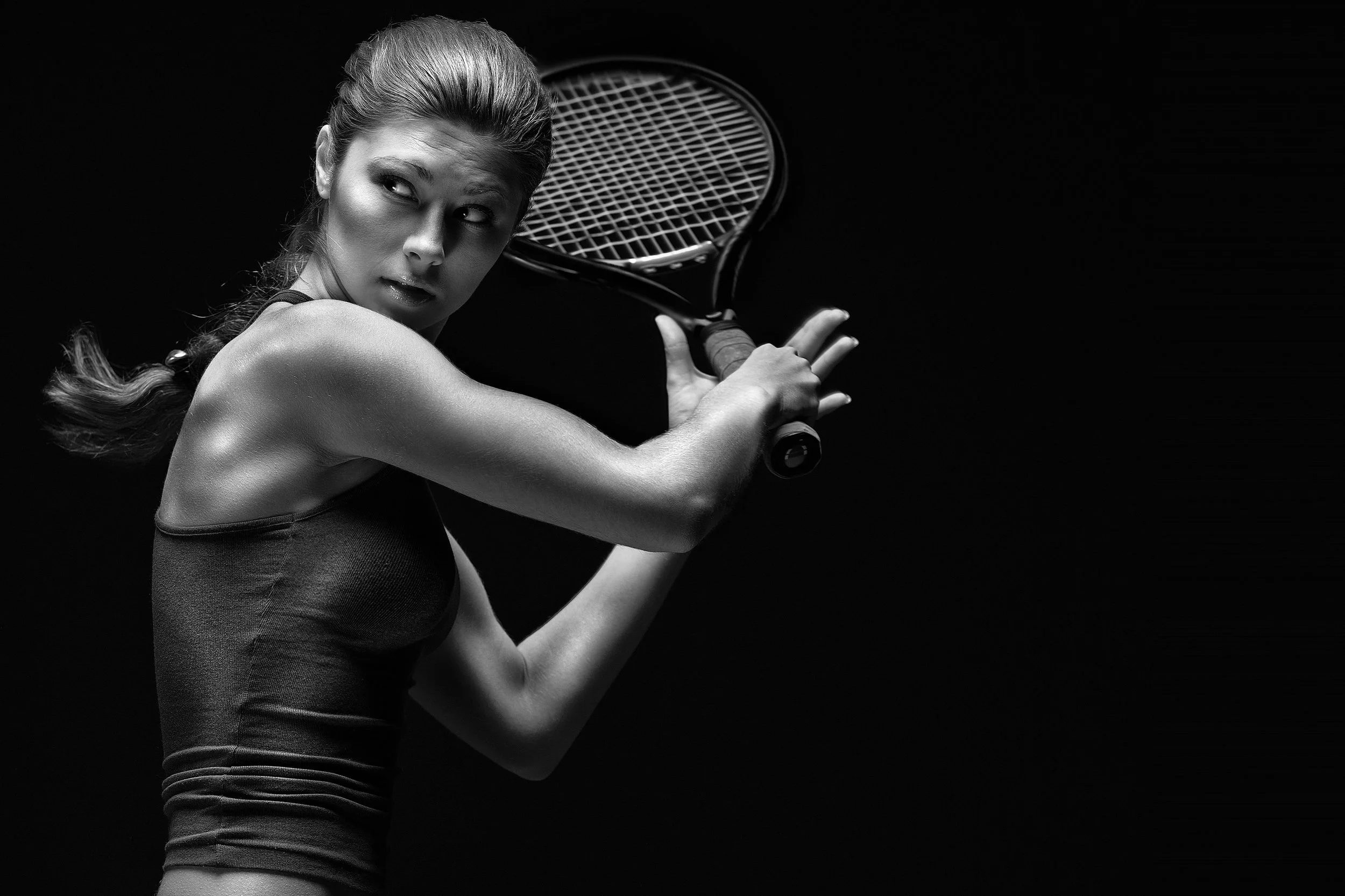 A woman with a ponytail holding a tennis racket in a ready position, against a black background.