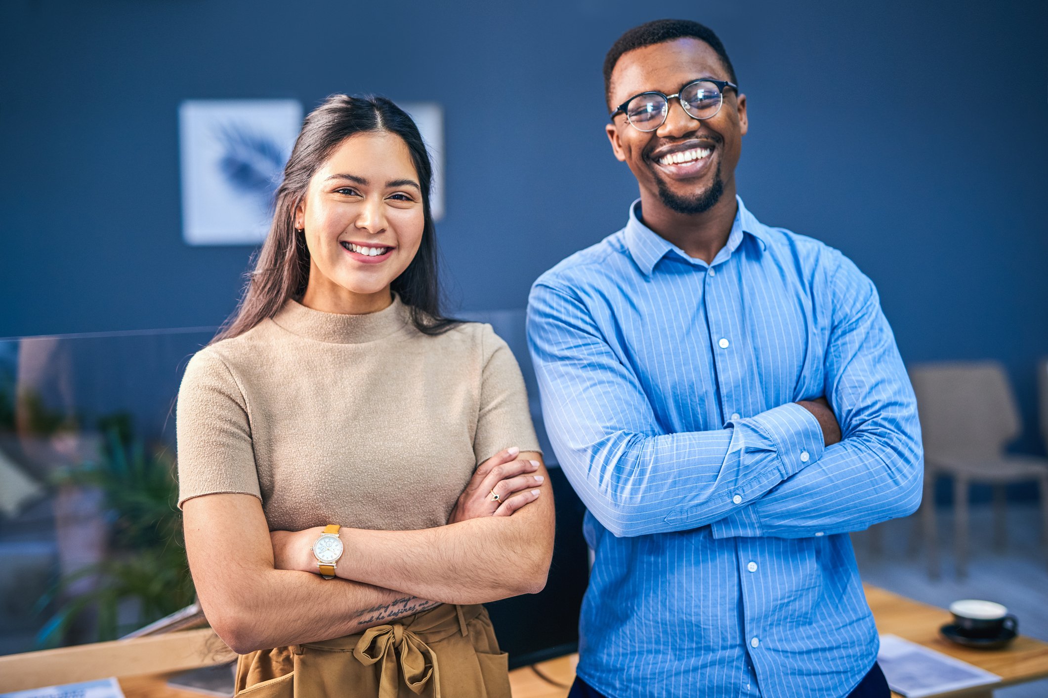 Two smiling professionals, a woman with long dark hair in a beige top and a man with glasses and a blue striped shirt, standing with arms crossed in a modern office setting.