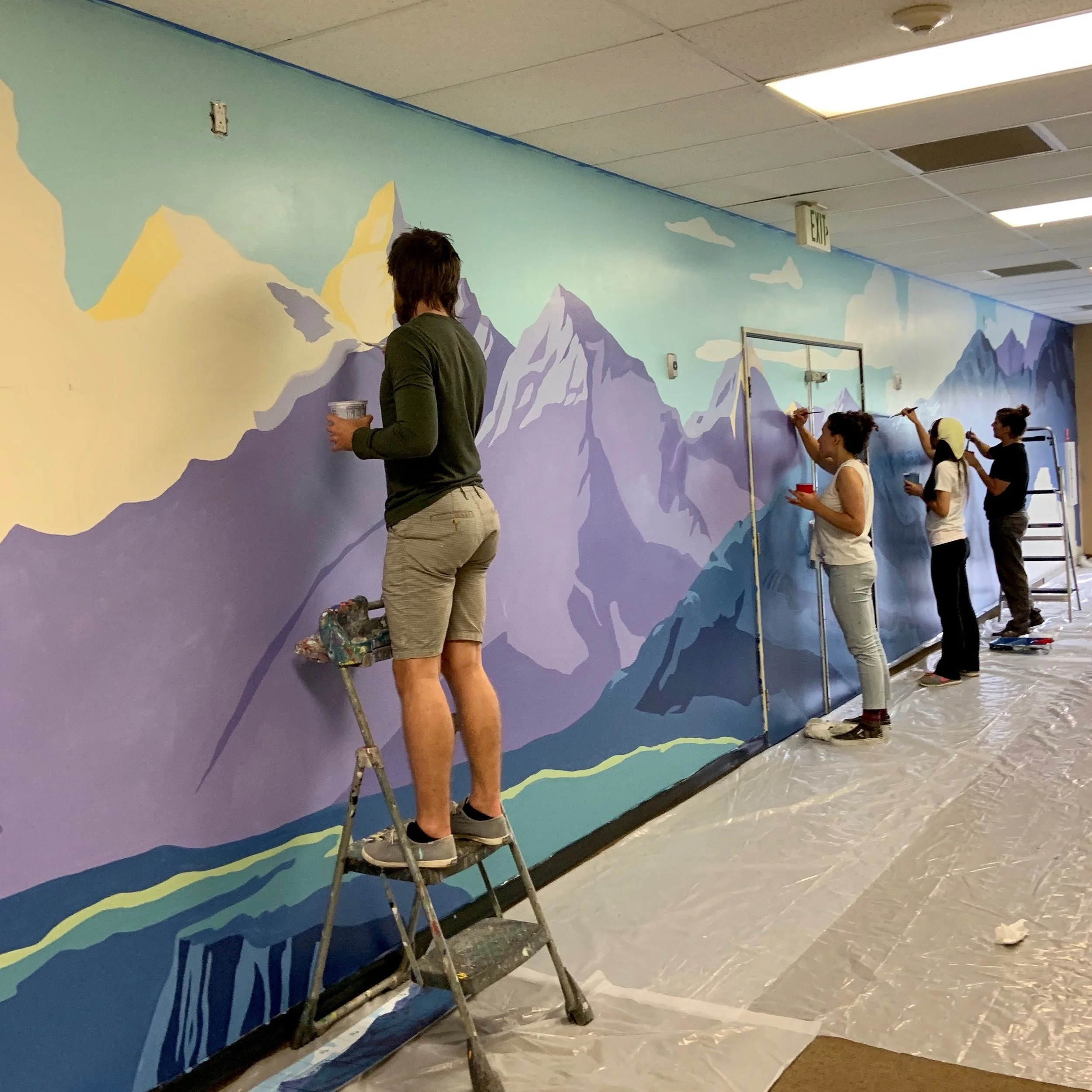 A group of four people painting a mural of mountain landscape on a wall inside a building. They are working on different sections of the mural, which features mountains, clouds, and a sky, using brushes and paints. A ladder and various painting suppl