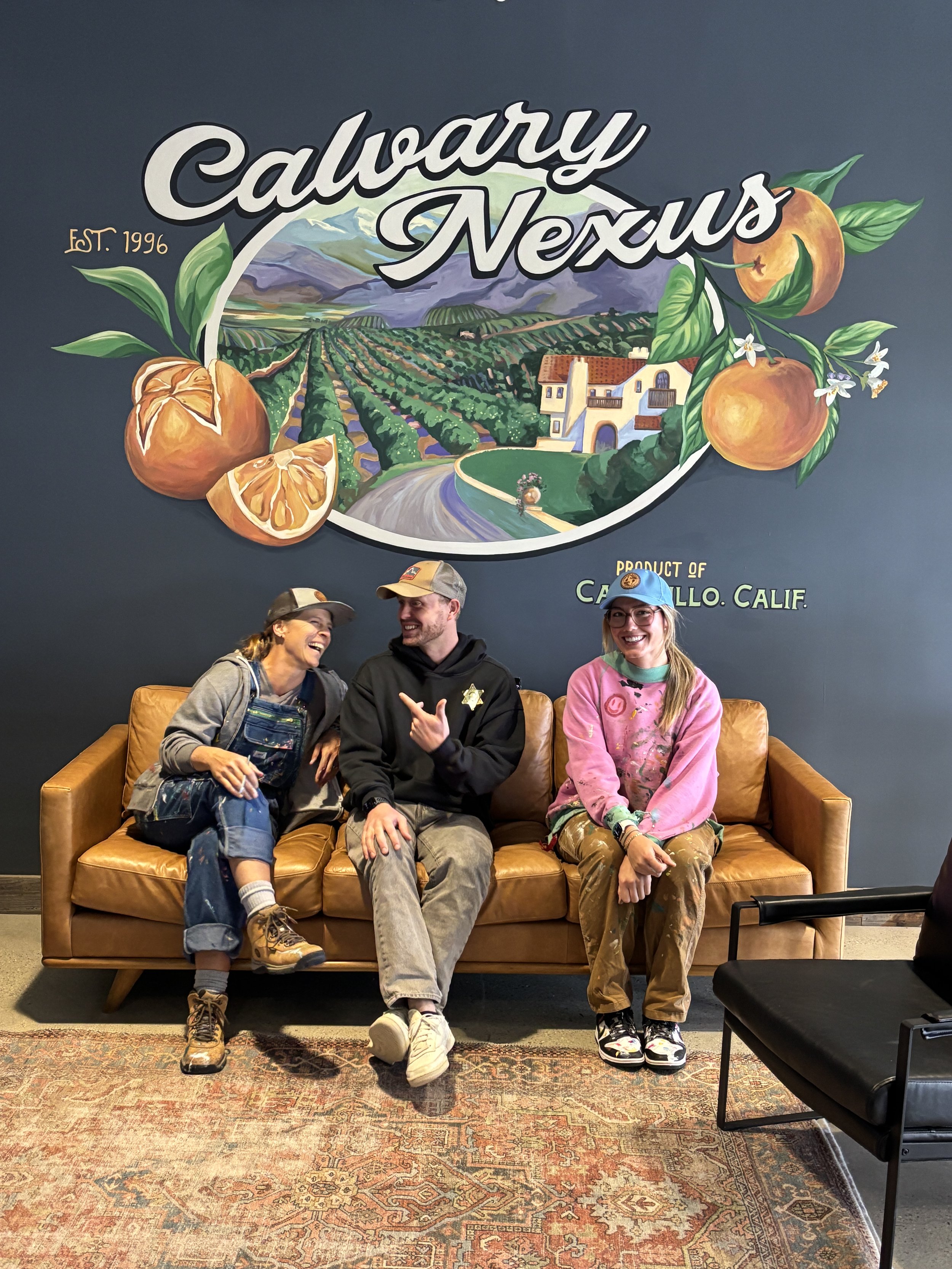 Three people sitting on a leather sofa in front of a mural that reads 'Calvary Nexus' with painted scenes of a vineyard and mountains. The person on the left is a woman with a hat, overalls, and boots, laughing. The man in the middle is wearing a cap