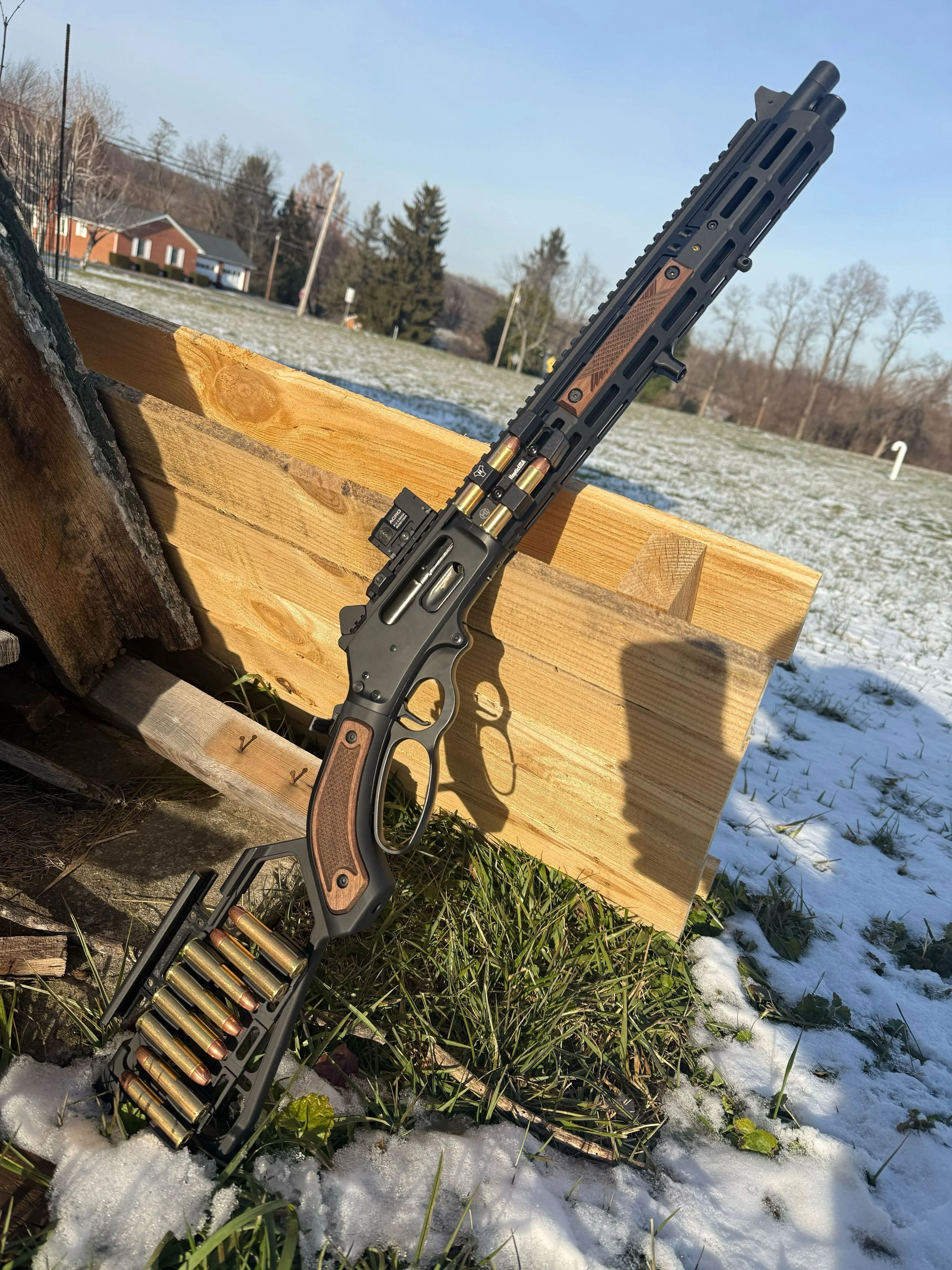 A black and wood finished lever-action rifle with a long barrel and attached magazine, leaning against a wooden fence outdoors in a snowy field.