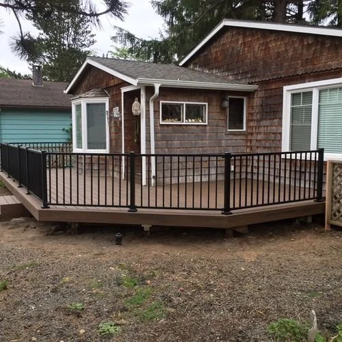 A newly built wooden deck with black metal railings attached to a house with brown shingle siding and white trim, with trees in the background.