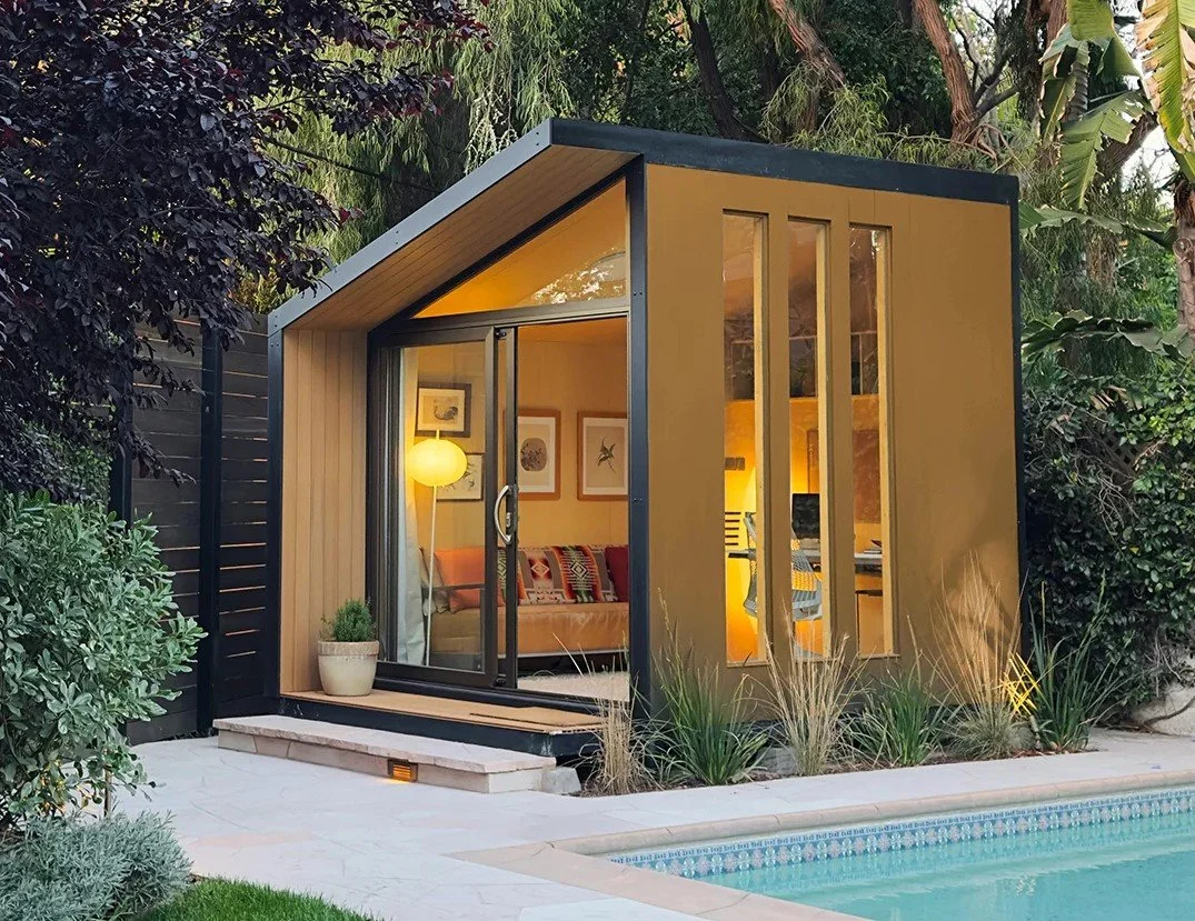 A modern backyard office pod with glass sliding doors and tall narrow windows, surrounded by plants and a swimming pool.