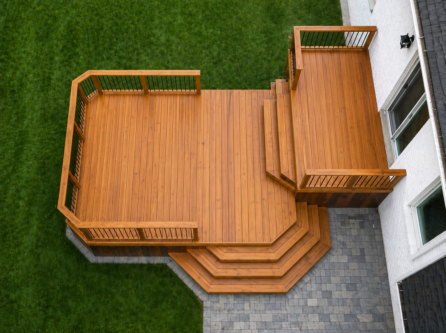 Overhead view of a wooden deck with stairs, attached to a white house, surrounded by green grass and a paved area.