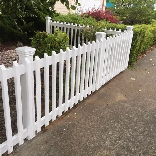 White picket fence along sidewalk with greenery and bushes in the background.
