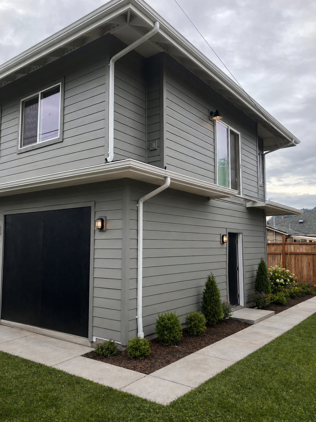 A modern two-story ADU with gray siding and black accents, surrounded by a small green lawn, landscaped bushes, and a concrete walkway. The house features a black garage door, several windows, and exterior lighting.