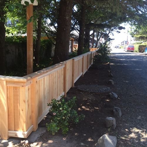New wooden fence with columns along a sidewalk in a residential area, with trees and parked cars in the background