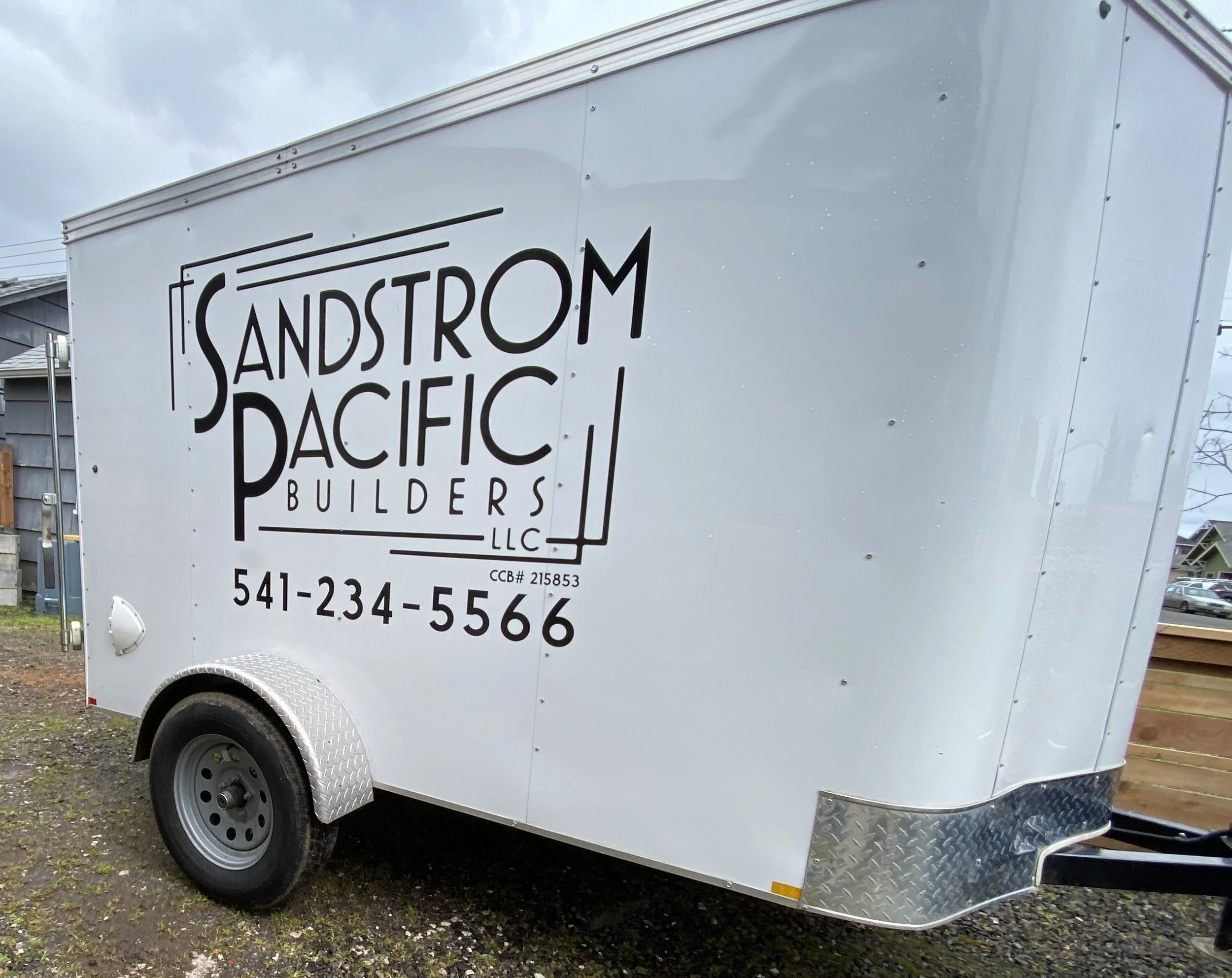A white cargo trailer with black lettering reading 'Sandstrom Pacific Builders LLC' parked on a gravel lot with a cloudy sky overhead.