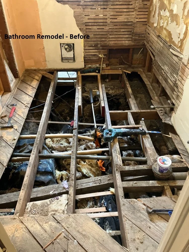 "Before" photo of older bathroom wall and floor removed for renovation, showing exposed wood lath, wooden floor joists, insulation, pipes, and tools.