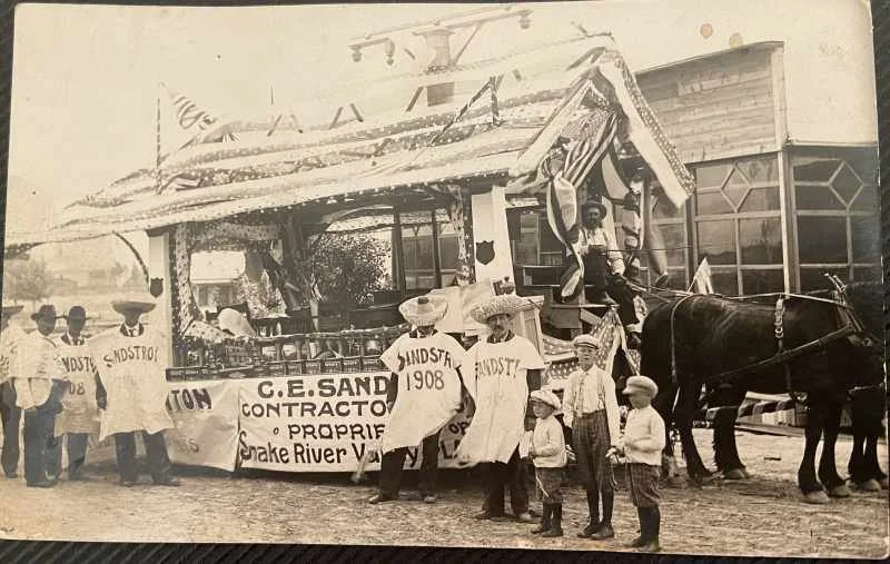 A vintage parade float decorated with banners and banners with people in old-fashioned clothing, some wearing hats, standing around. Two horses are hitched to the float, and a woman sits on it. The setting appears to be in front of a building with la