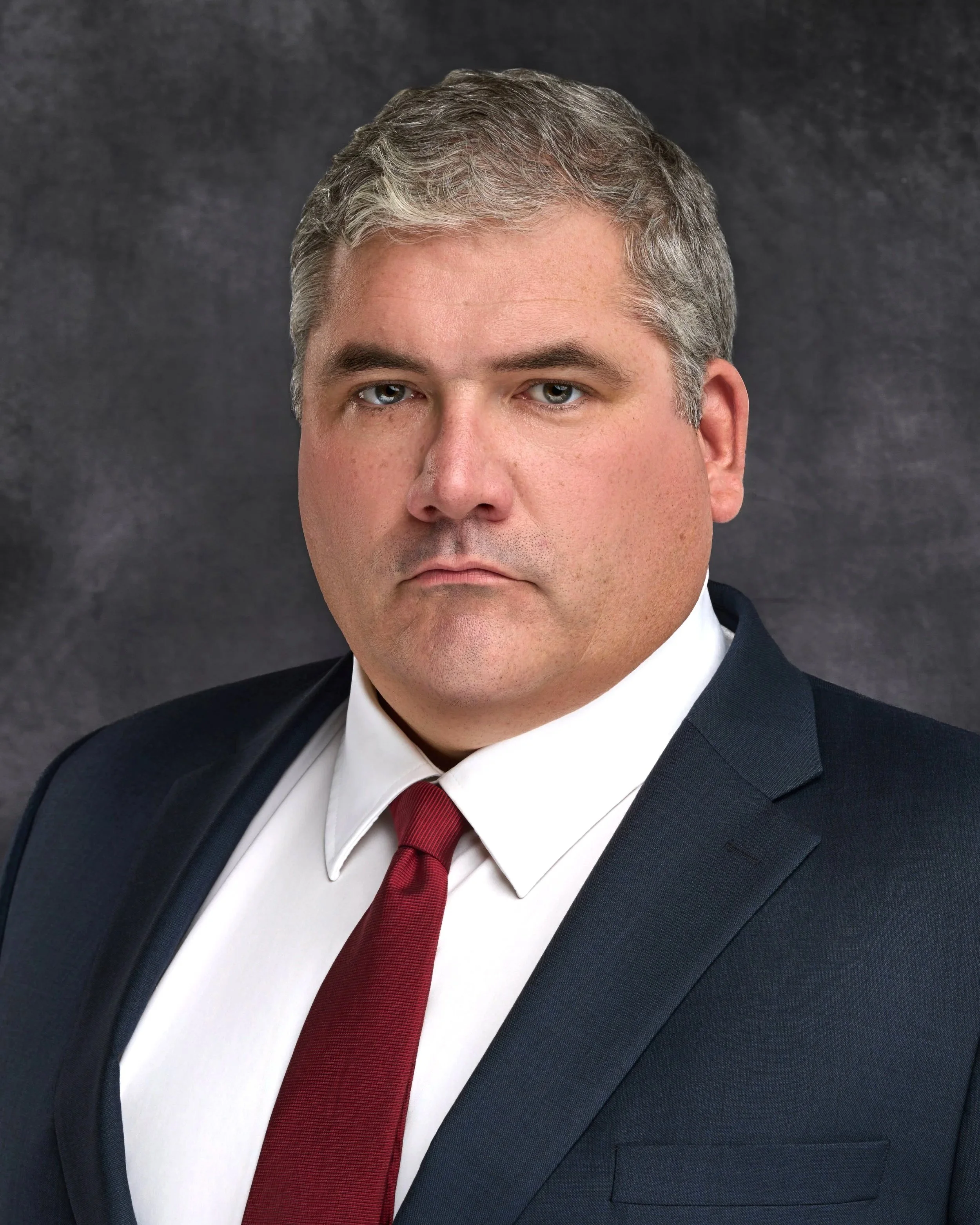 Chicago professional headshot portrait of a corporate man in a suit and red tie, with a serious expression, against a dark background.