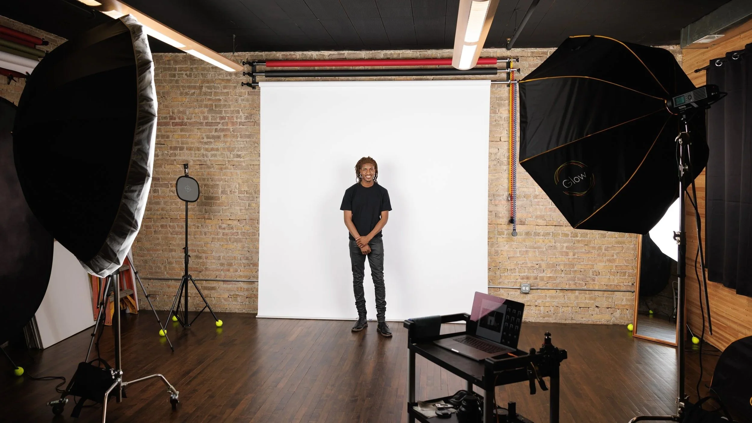 A behind-the-scenes portrait of a man taking modeling digitals standing in a photography studio with professional lighting equipment, a white backdrop, and a brick wall behind him.