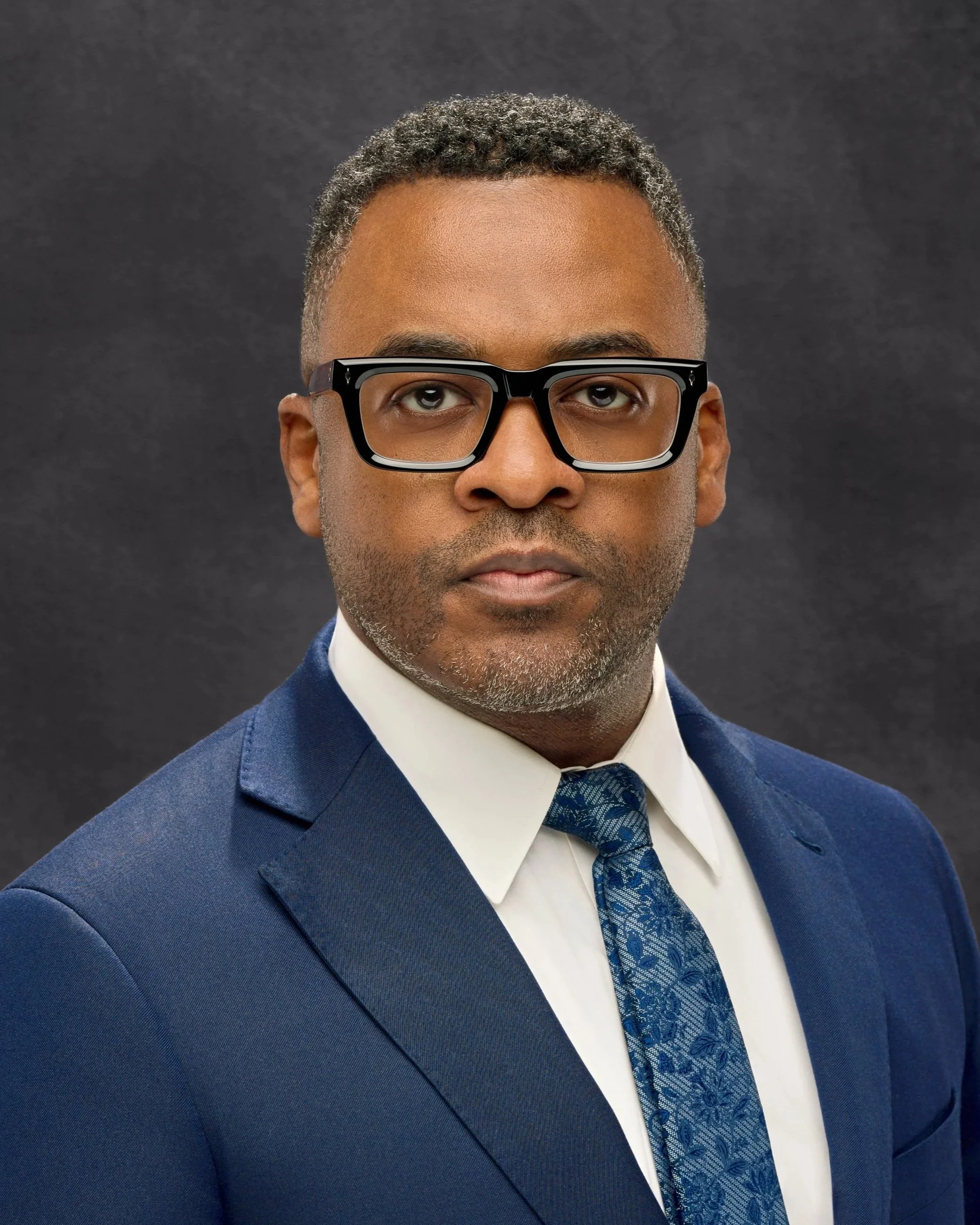 Chicago professional headshot portrait of business man in a blue suit, white shirt, and patterned blue tie, posing against a dark background.