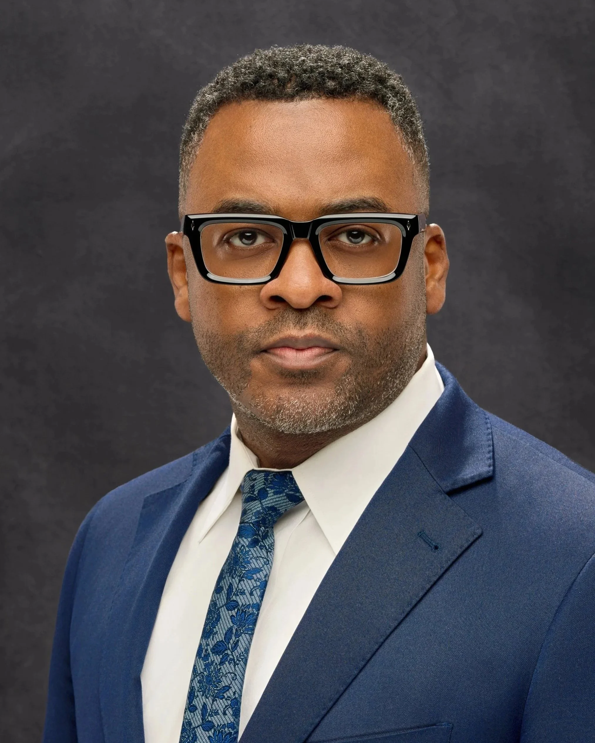 Chicago professional headshot portrait of business man in a navy suit, white shirt, and patterned blue tie, wearing black glasses, against a dark background.