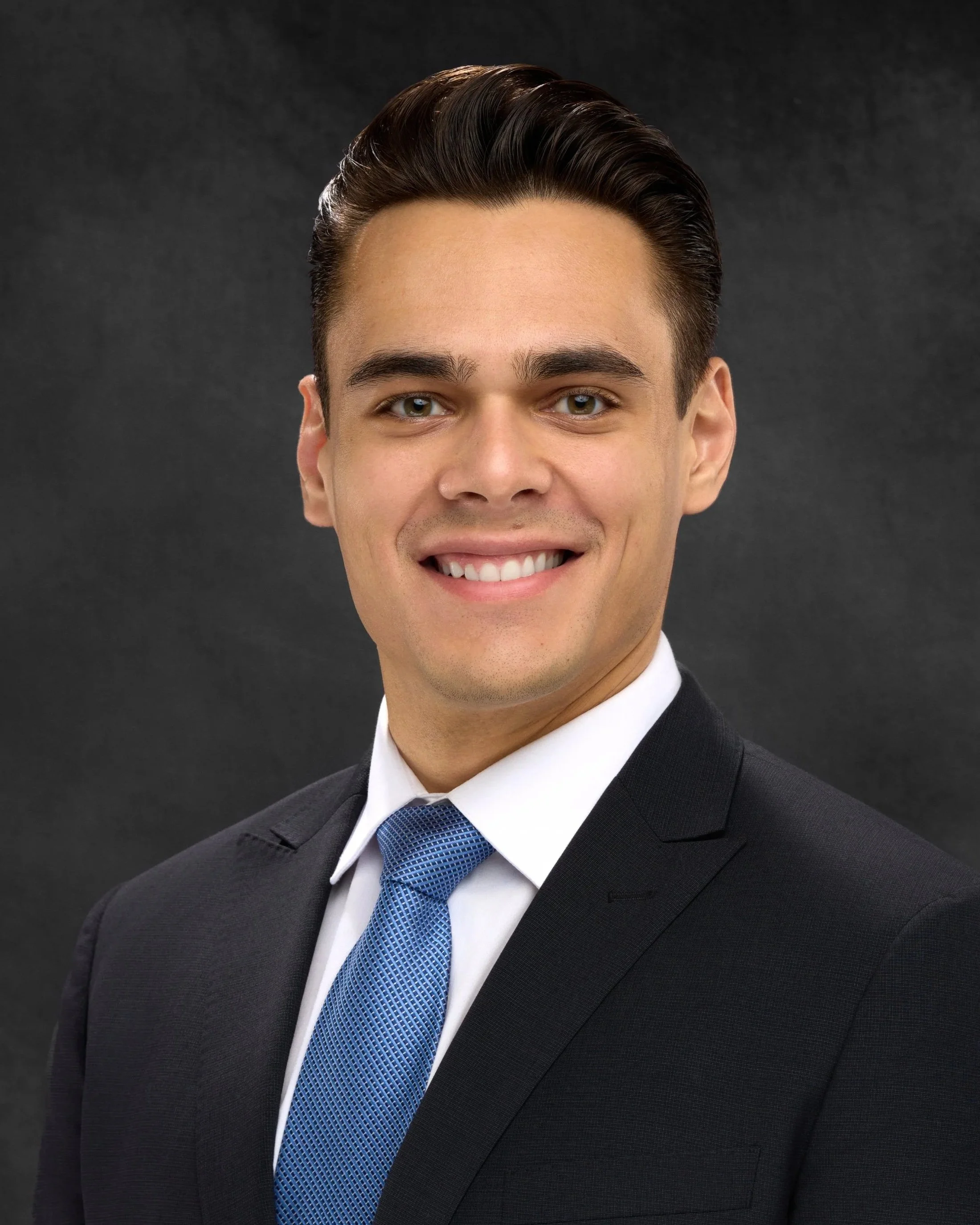 Chicago professional headshot portrait of business man with dark hair styled back, smiling, dressed in a black suit, white shirt, and blue tie, against a dark gray background.