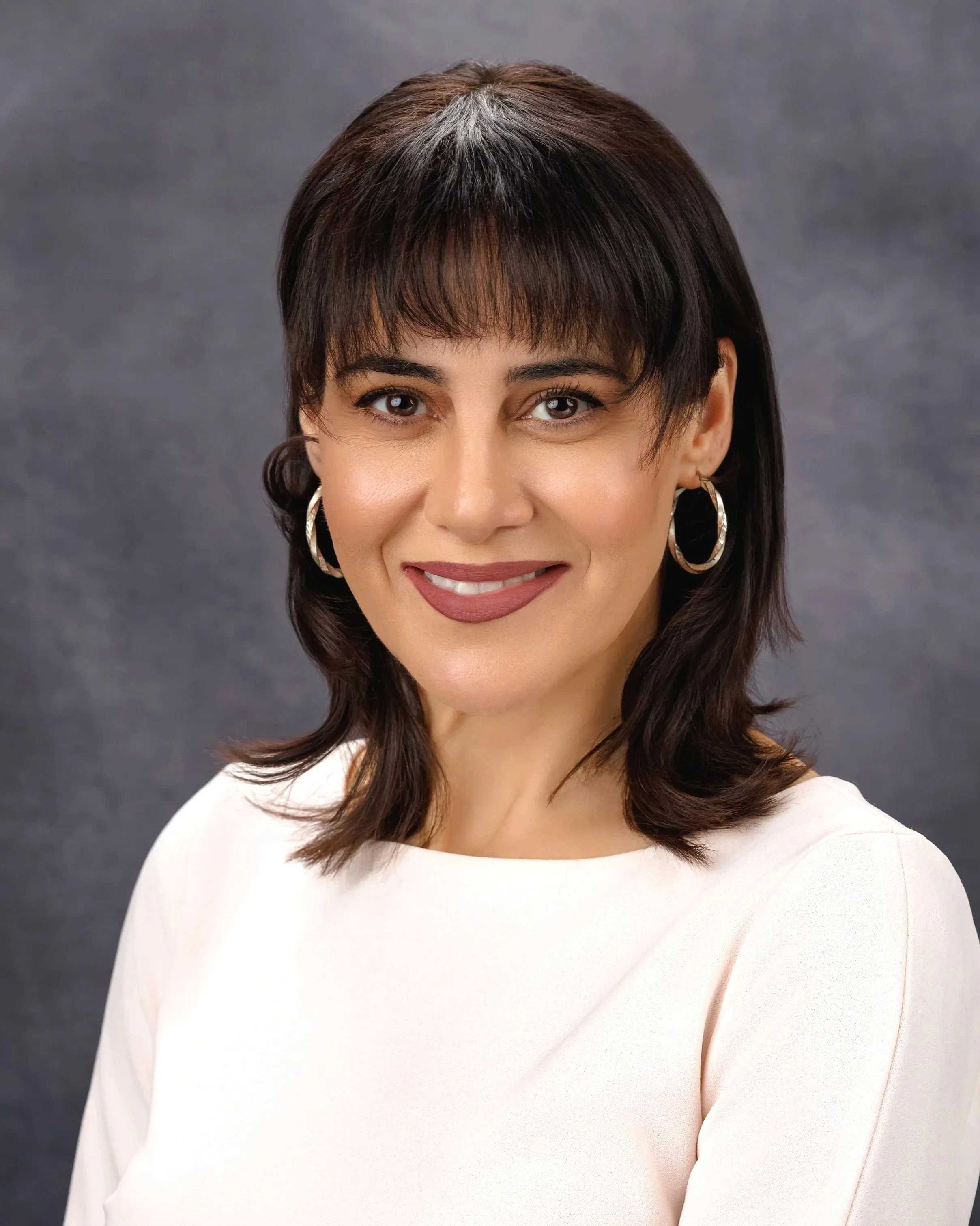 Chicago professional headshot portrait of a female real estate agent with dark brown hair, wearing hoop earrings and a white top, smiling in front of a gray background.