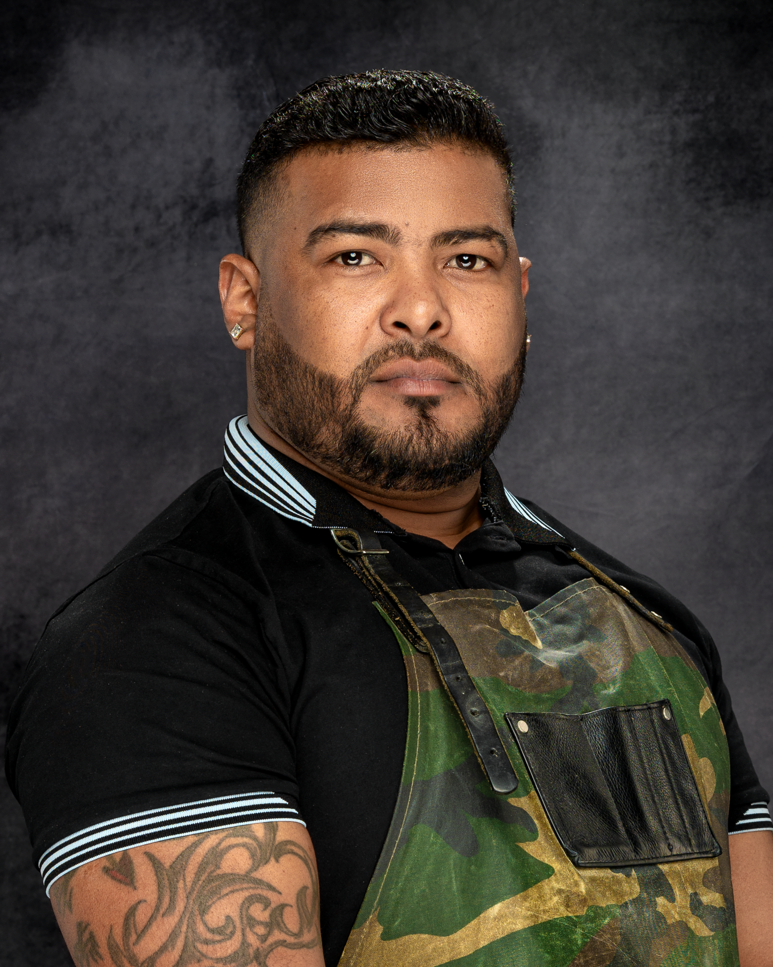 Professional branding headshot portrait of a man with a beard and short dark hair wearing a black shirt with a white striped collar and sleeves, and a camouflage apron, stands against a dark textured background.