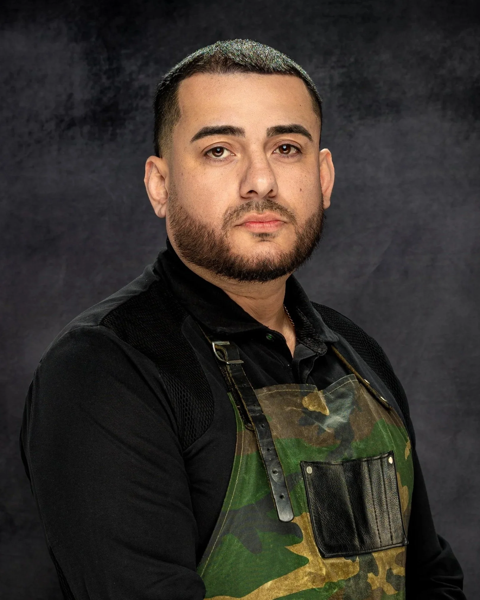 Professional branding headshot portrait of a man with short dark hair, beard, and mustache wearing a black shirt and camouflage vest against a dark background.