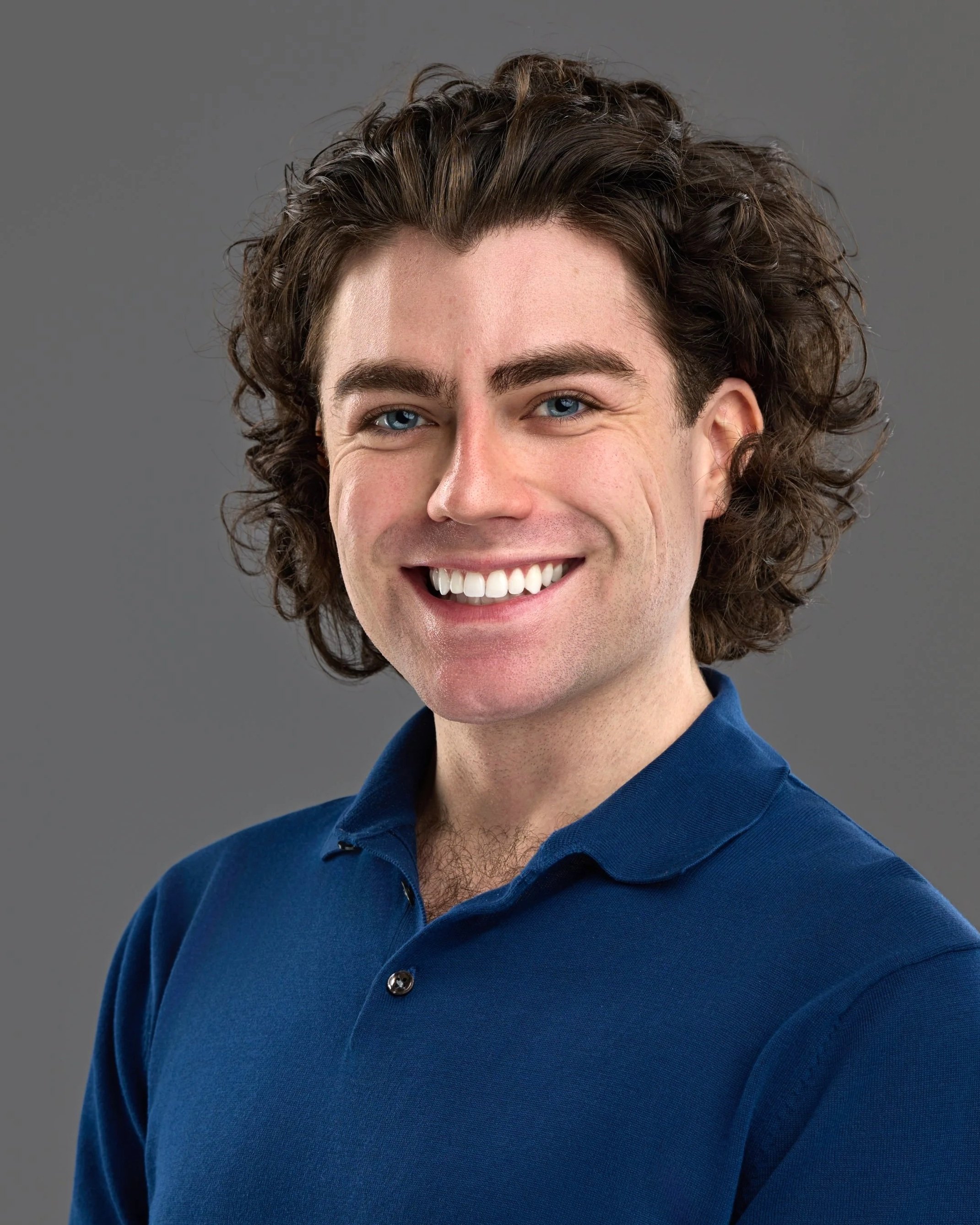 Chicago professional headshot portrait of a young actor with curly brown hair and blue eyes, smiling, wearing a blue polo shirt against a gray background.