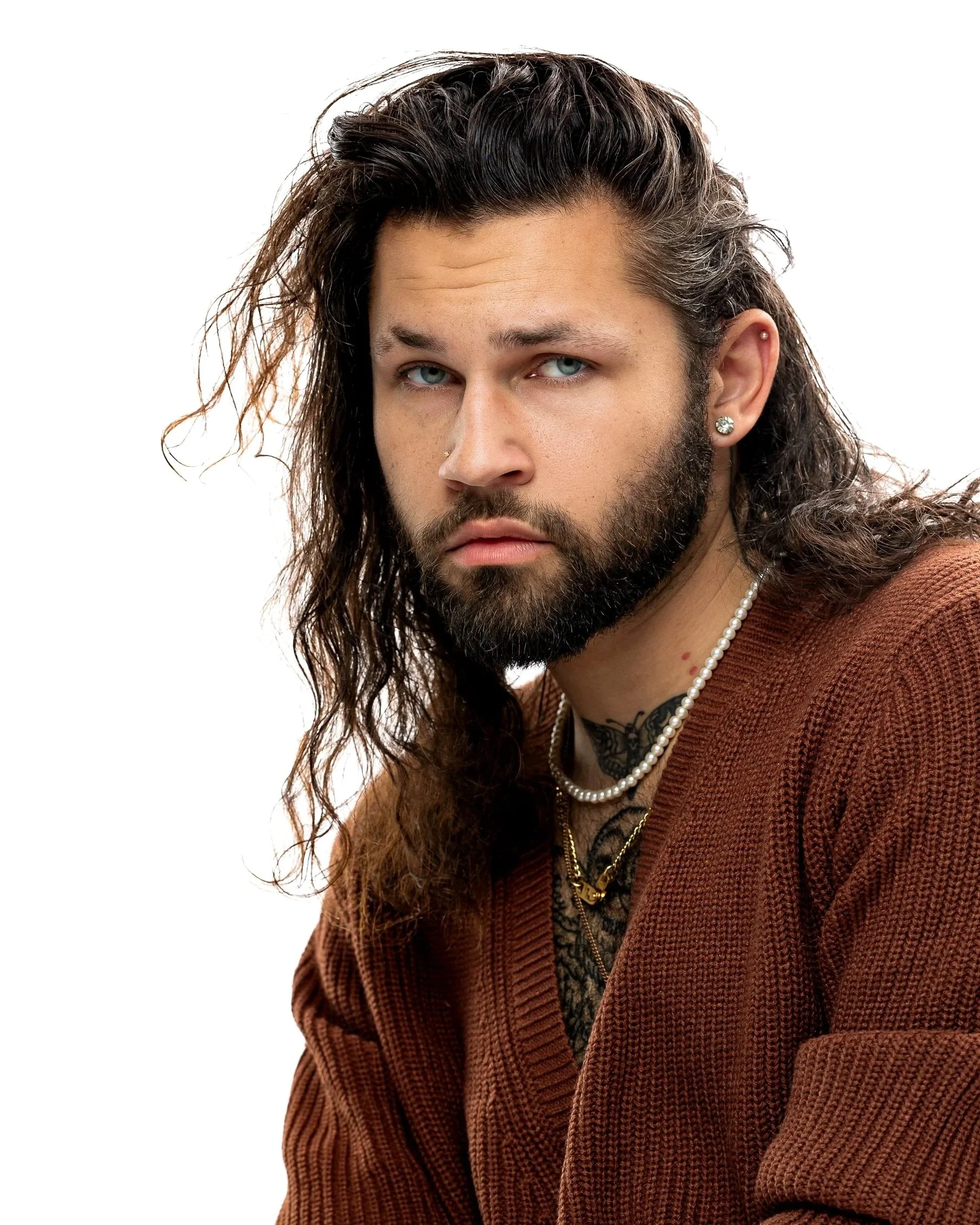 Chicago professional headshot portrait of an actor with long, wavy brown hair, blue eyes, and a beard, wearing jewelry and a brown shirt, against a white background.