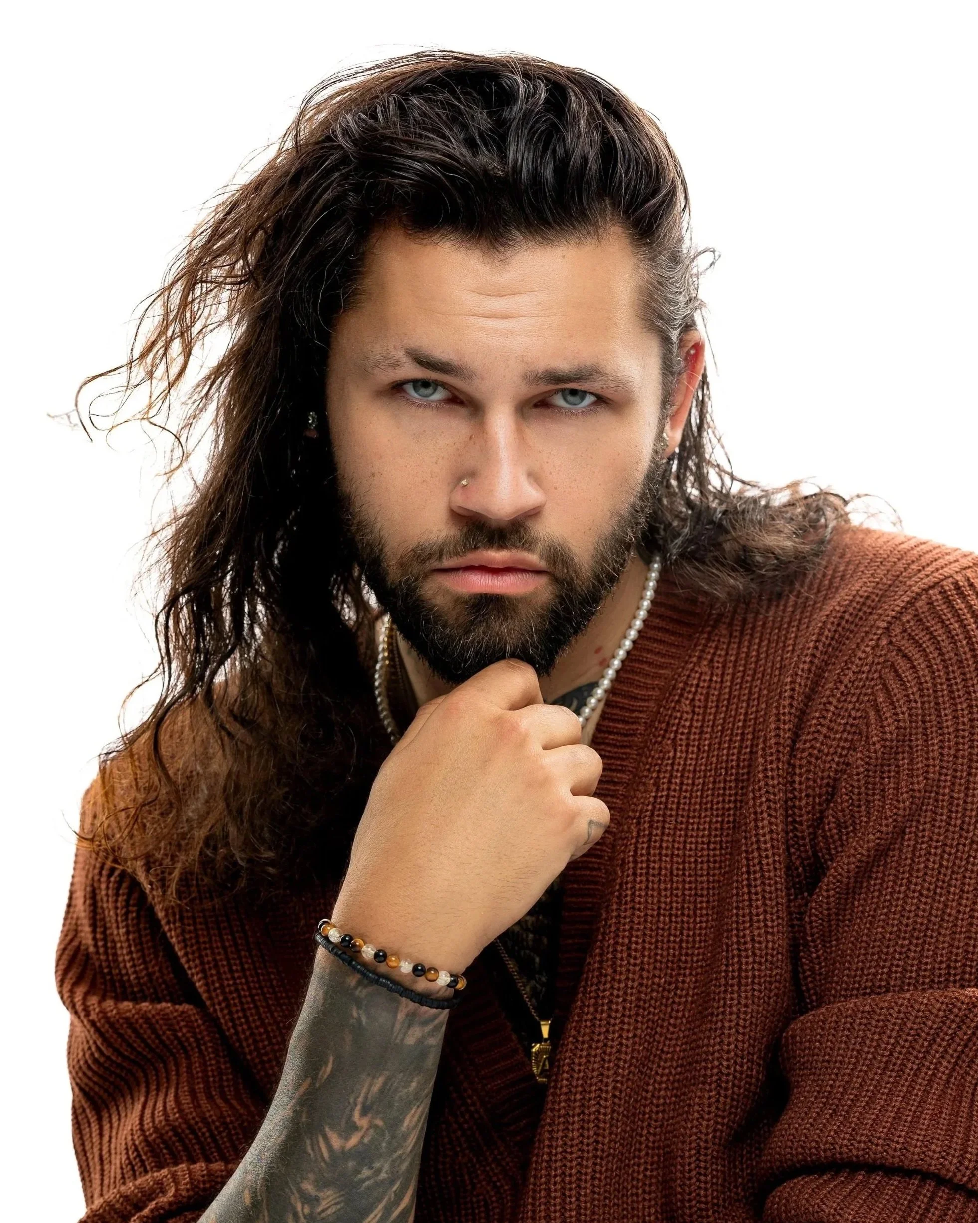 A young man with long, wavy dark hair and a beard looks intensely into the camera, resting his chin on his hand, wearing a brown sweater and accessories like a pearl necklace, beaded bracelet, and earrings.