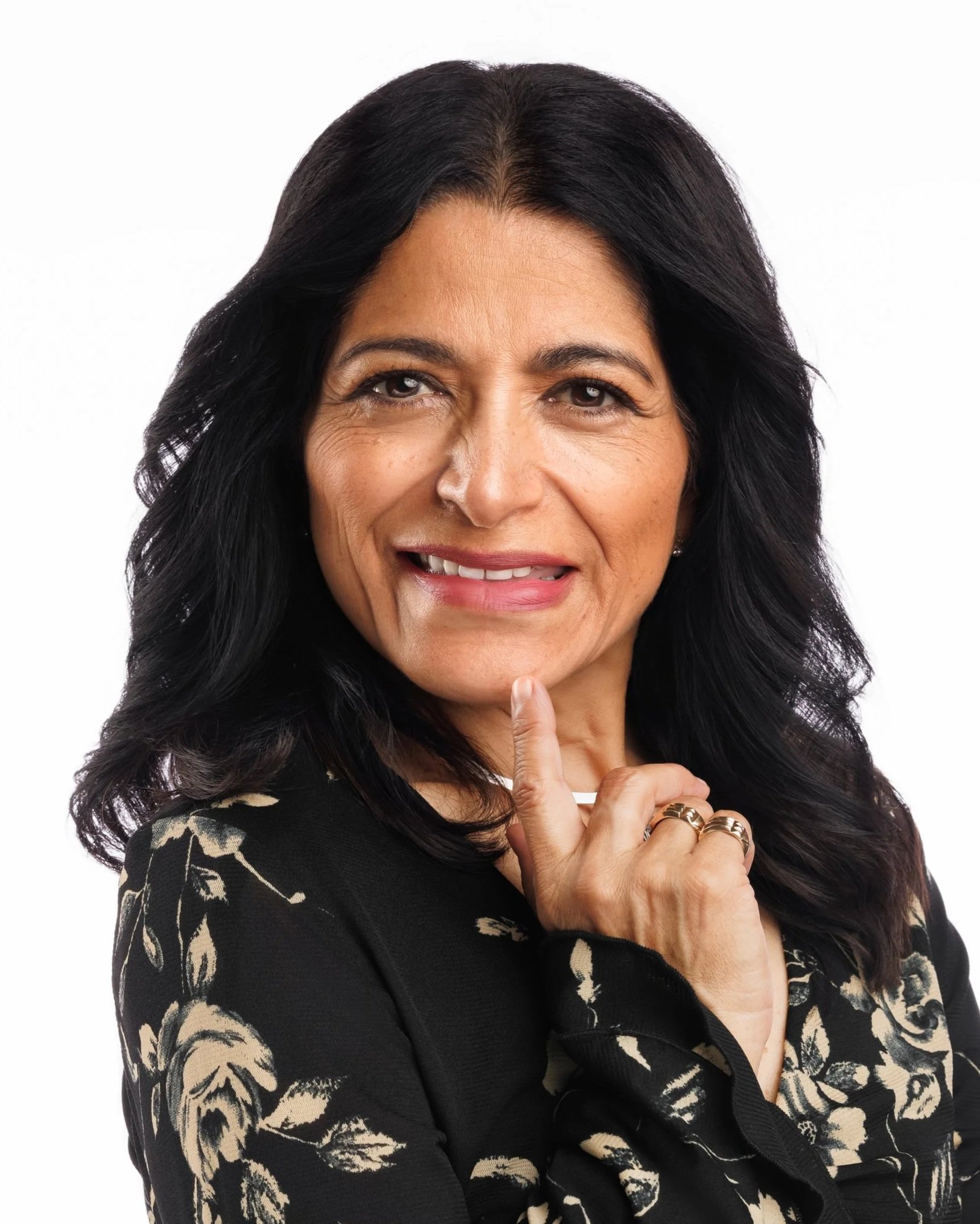 Professional branding headshot portrait of a middle-aged woman with dark wavy hair, making a playful expression with her tongue slightly out, touching her chin with her index finger, against a plain white background.