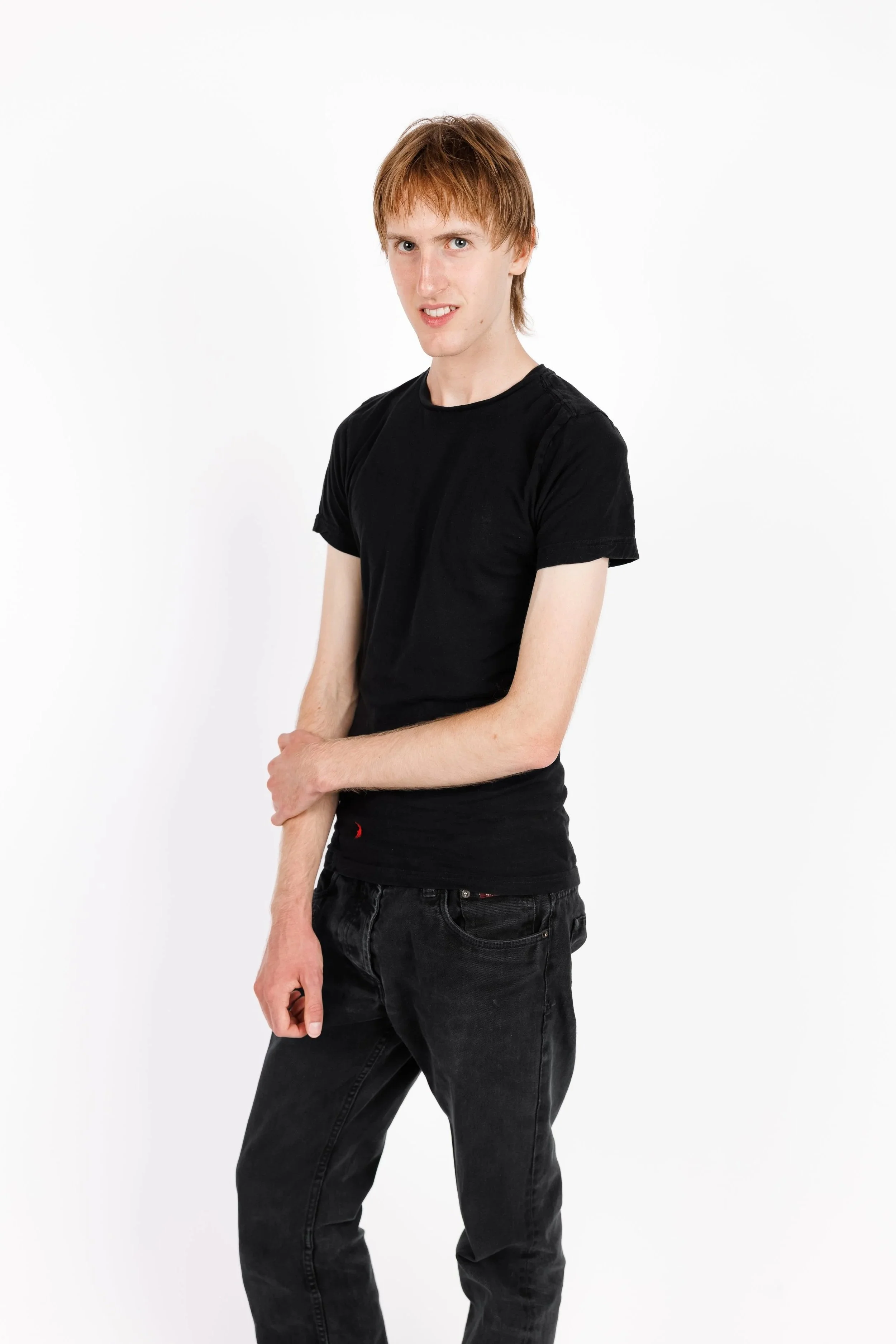 A young man with red hair, wearing a black t-shirt and black jeans, standing against a white background.