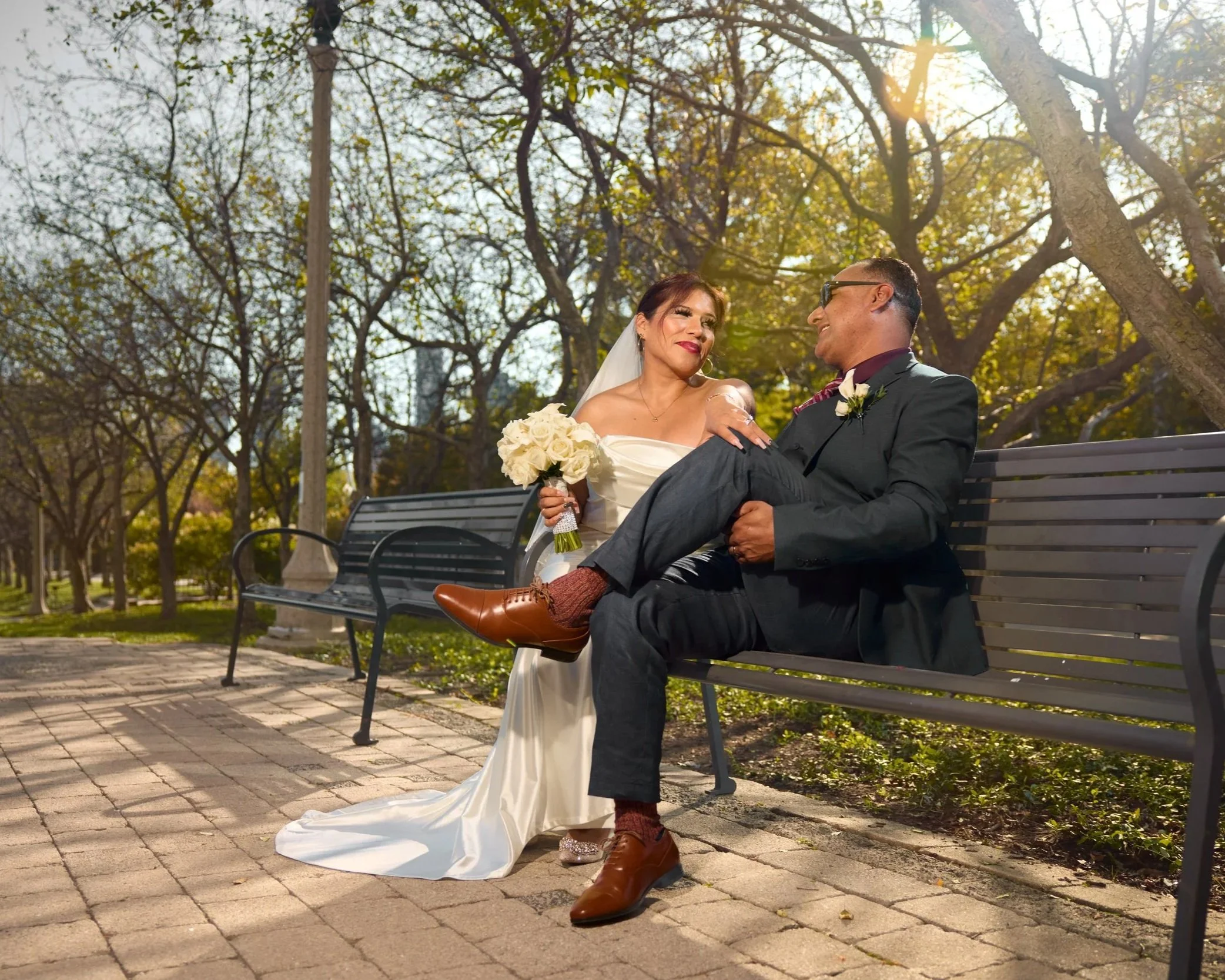 Bride in a white wedding dress and groom in a black suit sitting on a park bench, surrounded by trees with sunlight filtering through, during a wedding photoshoot.