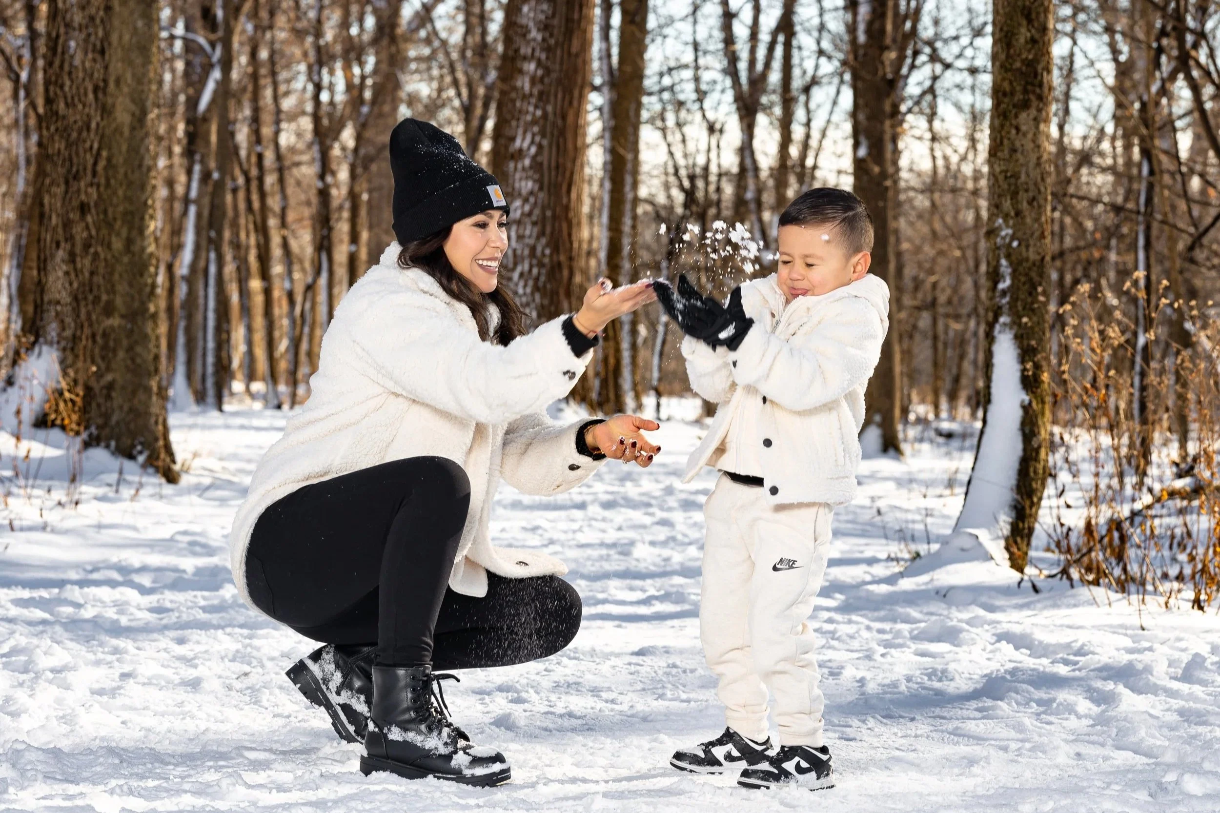 A woman and young boy in white winter clothing playing in a snowy forest, with the woman squatting and the boy showing a playful expression as they enjoy snow activity.