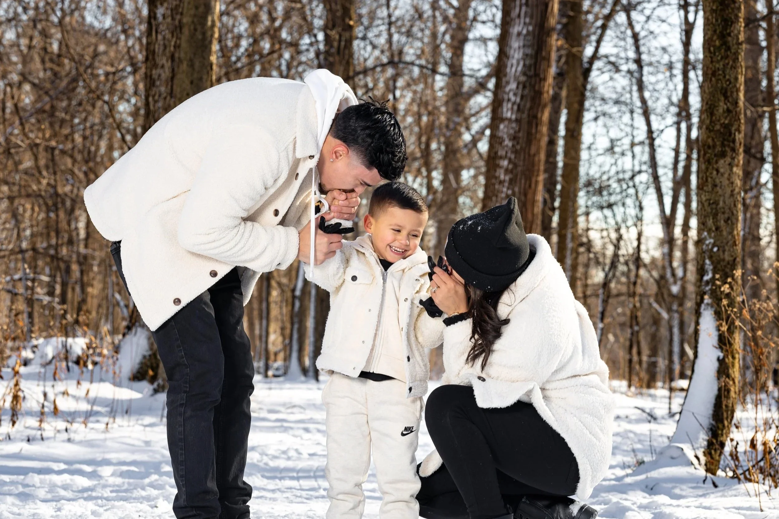 A family of three in winter clothing, smiling and playing together in a snowy forest while taking photographs of a young boy.