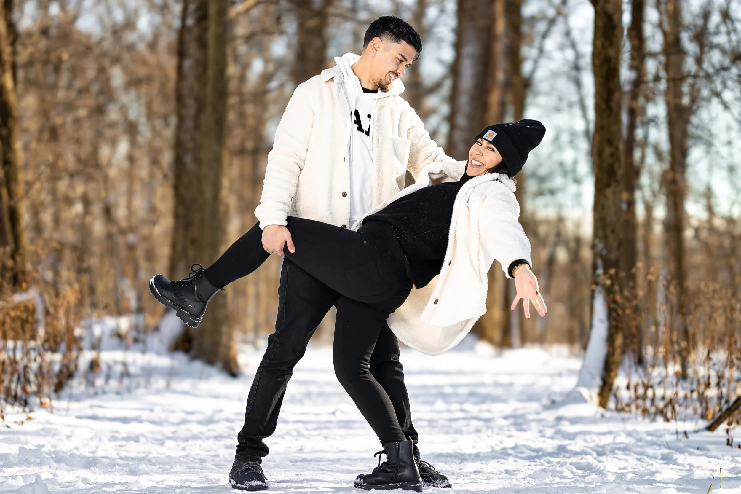 A couple enjoying snowy day outdoors, smiling, with the man lifting the woman in a snow-covered forest.