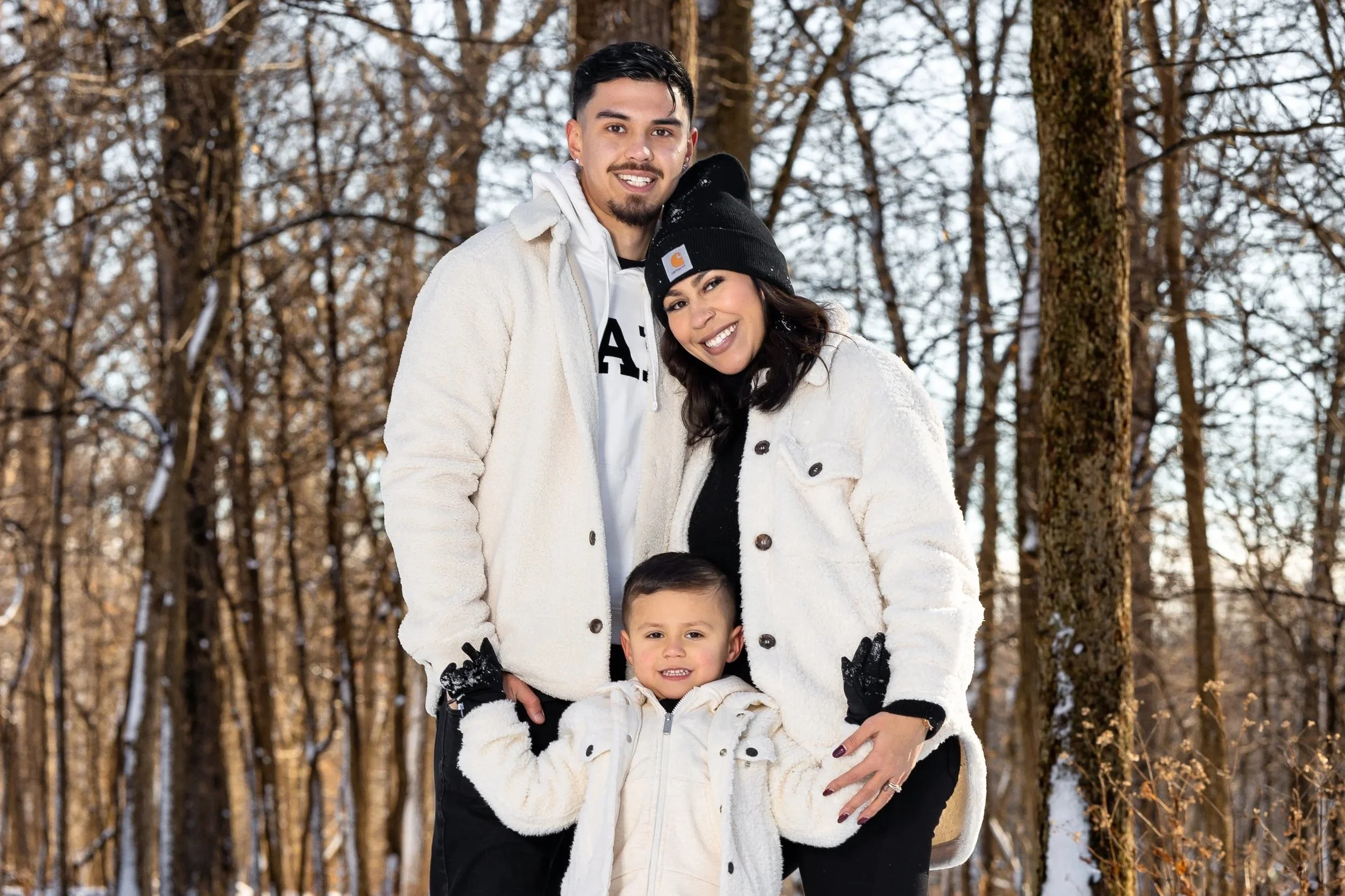 Family of three standing in a snowy forest, smiling and bundled up in warm, white coats and black gloves.