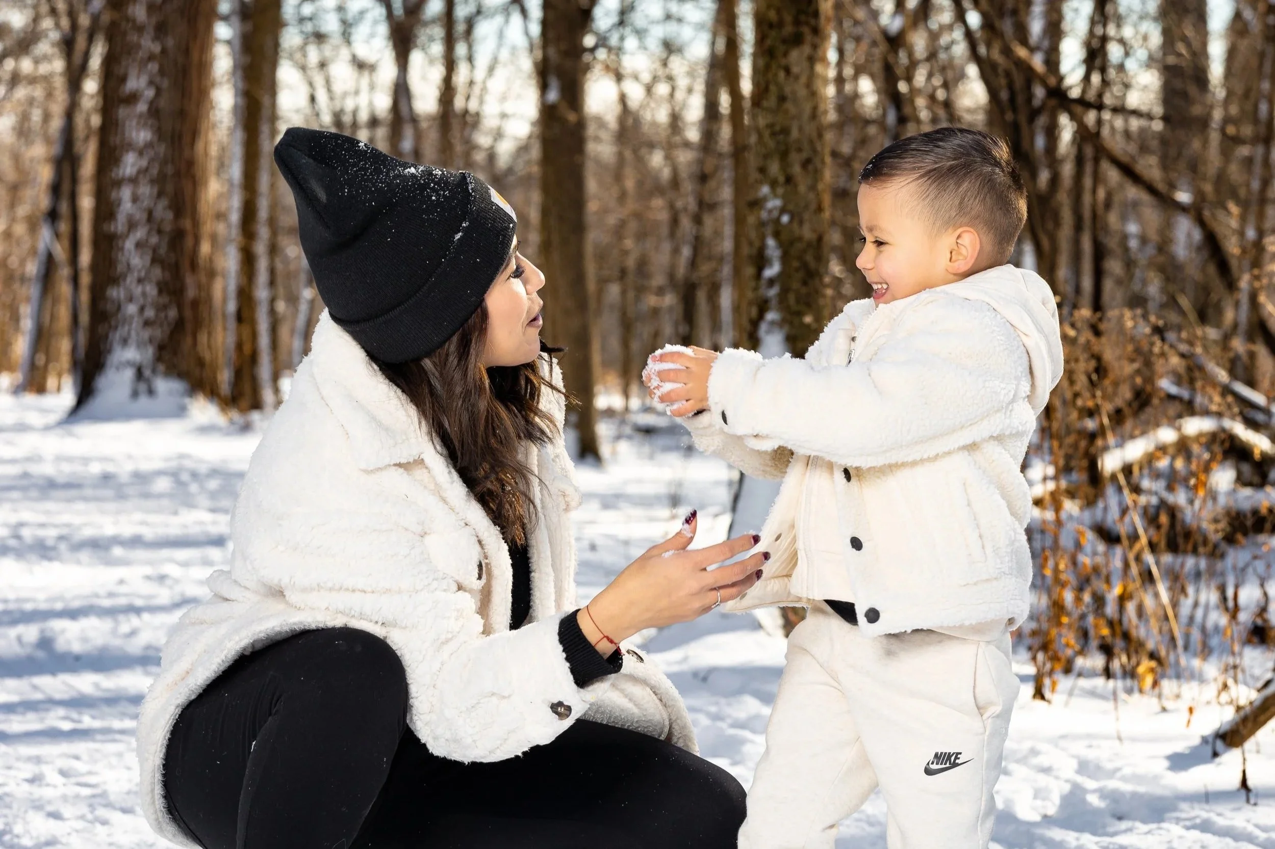 A woman and a young boy playing in a snowy forest, both dressed in winter clothing. The woman is crouched down, facing the boy, who is smiling and holding snow in his hands.