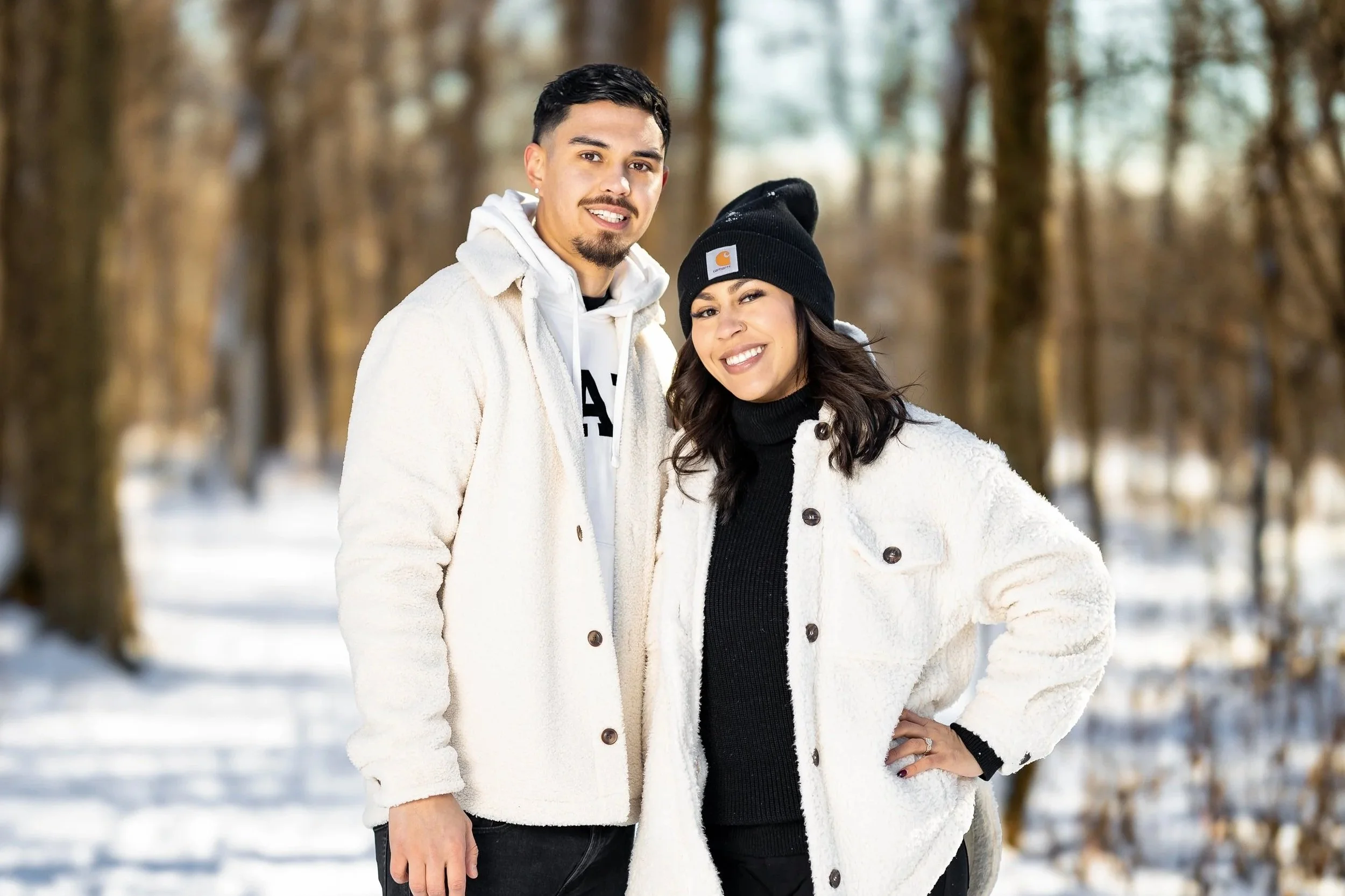 A smiling young man and woman standing close together in a snowy forest, dressed warmly in white and black winter clothing.