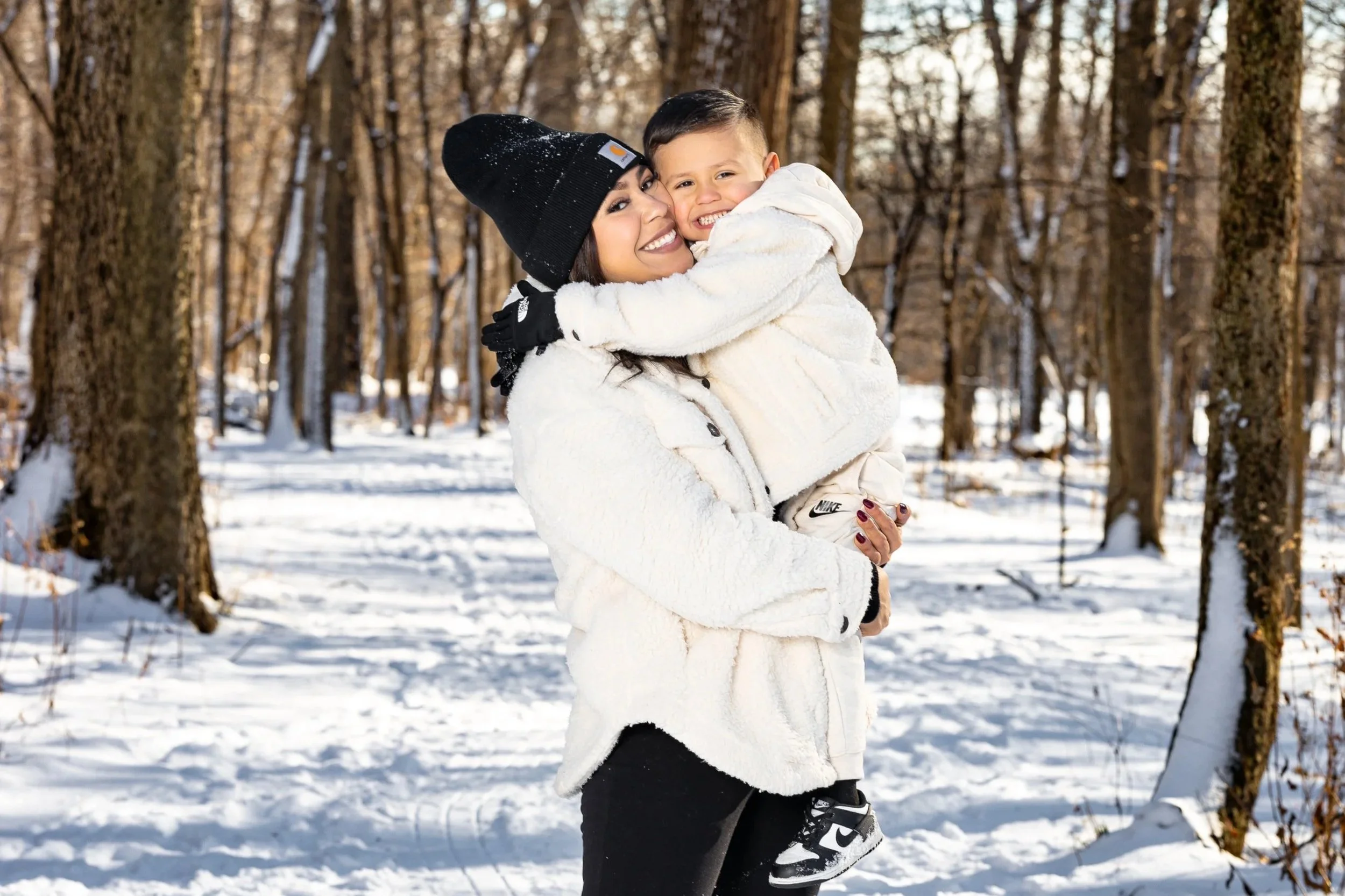 A woman and a young boy enjoying a snowy forest, with the woman holding the boy and both smiling happily at the camera.