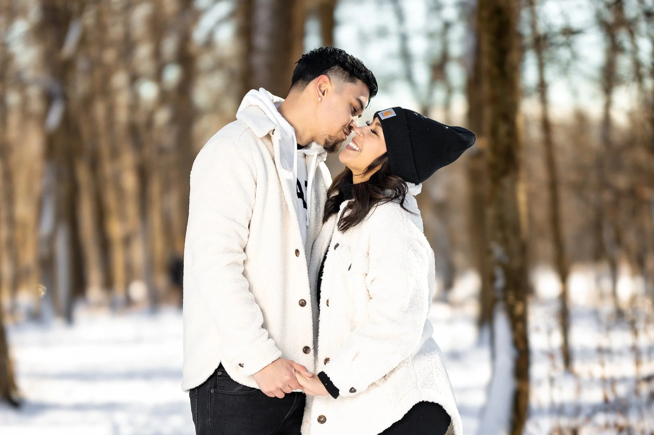 A smiling couple standing close in a snowy forest, touching foreheads and holding hands, both dressed in warm winter clothing.