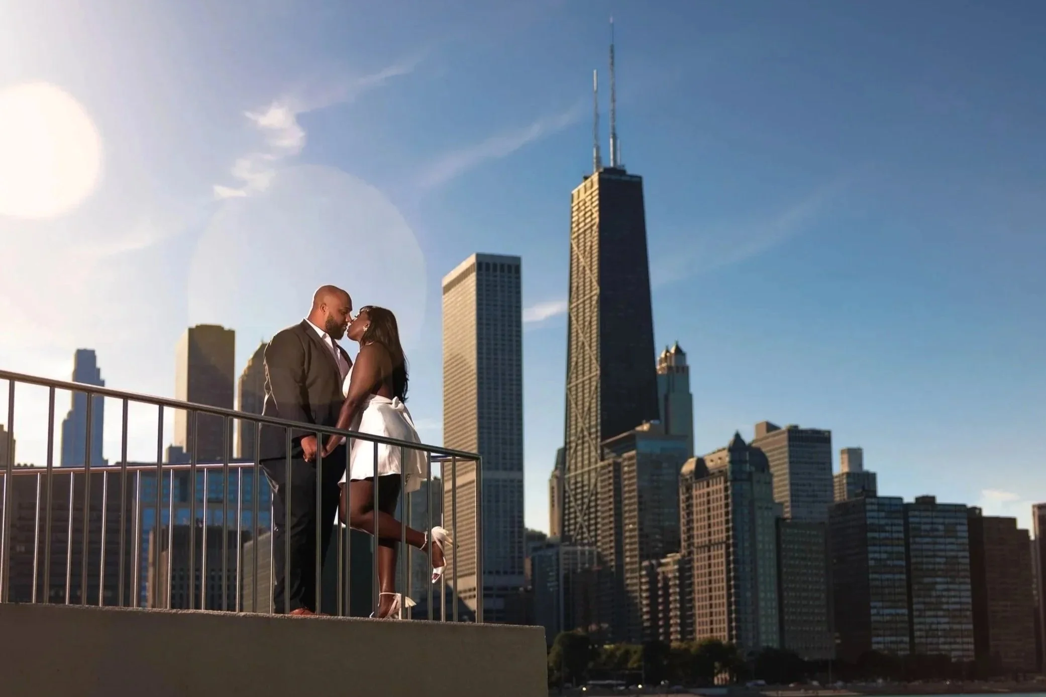 A couple dressed in formal attire standing on a balcony over a body of water with the Chicago skyline in the background during sunset.