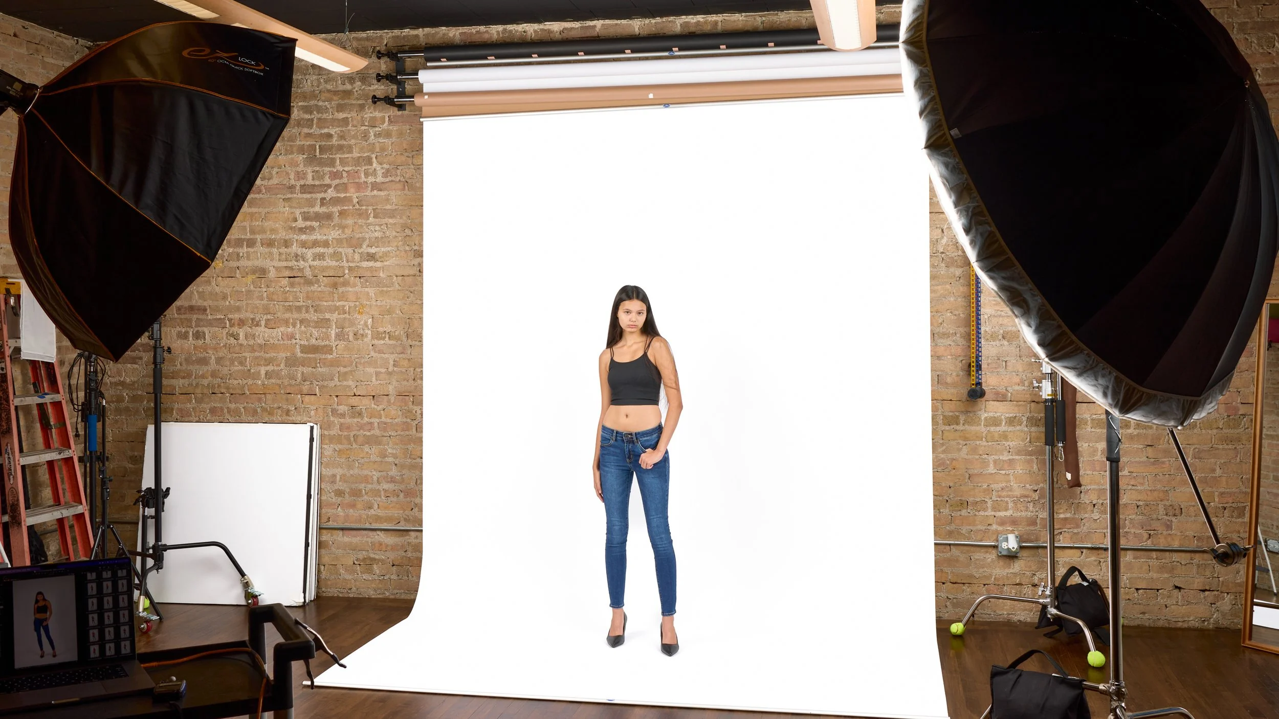A behind-the-scenes portrait of a woman in a studio posing in a photography studio with a plain white backdrop, professional lighting equipment on either side, and a brick wall background.