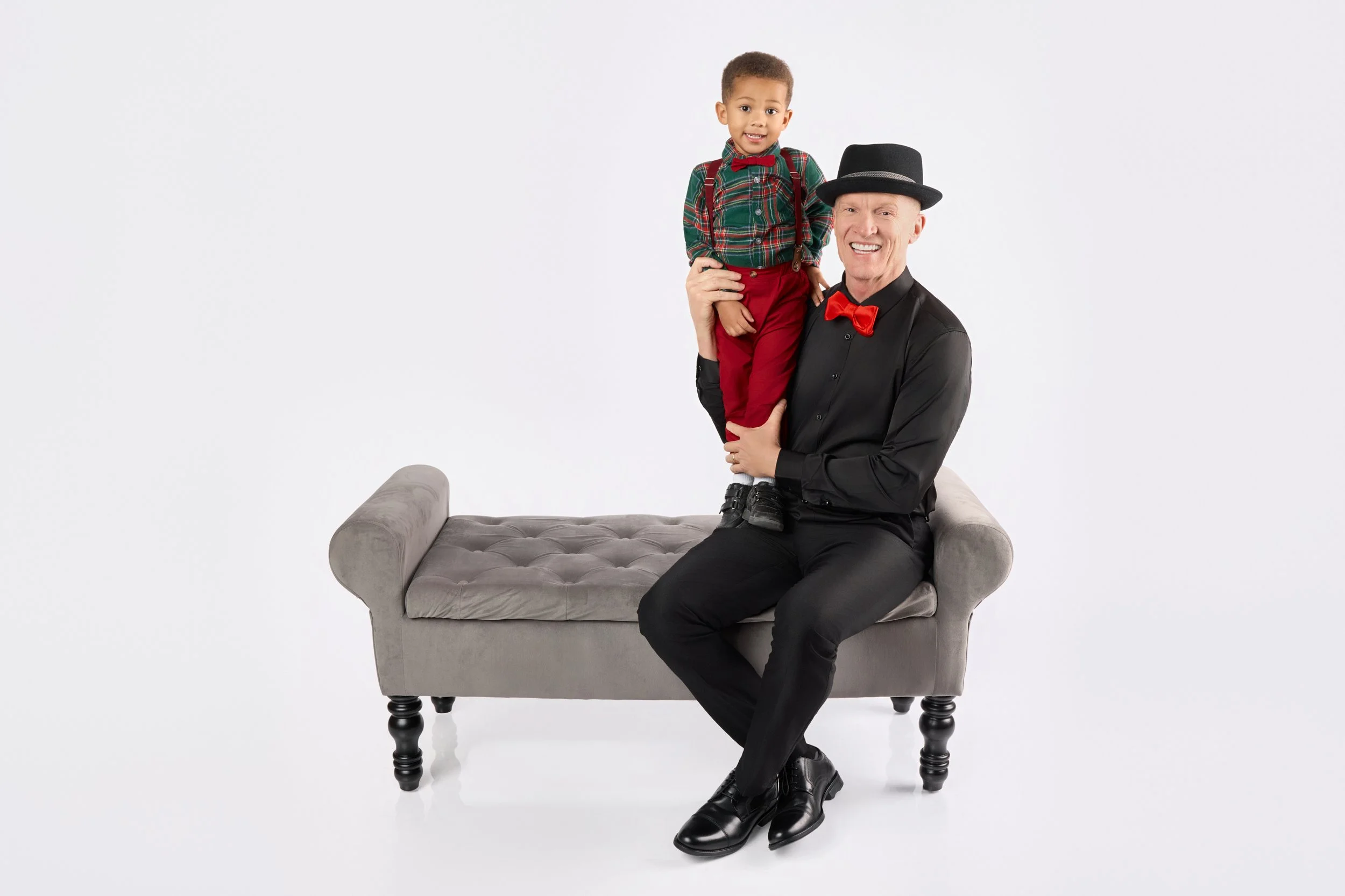 A man dressed in black with a red bow tie and black hat sitting on a gray tufted bench, holding a young boy dressed in red and green plaid with red pants, against a plain white background.
