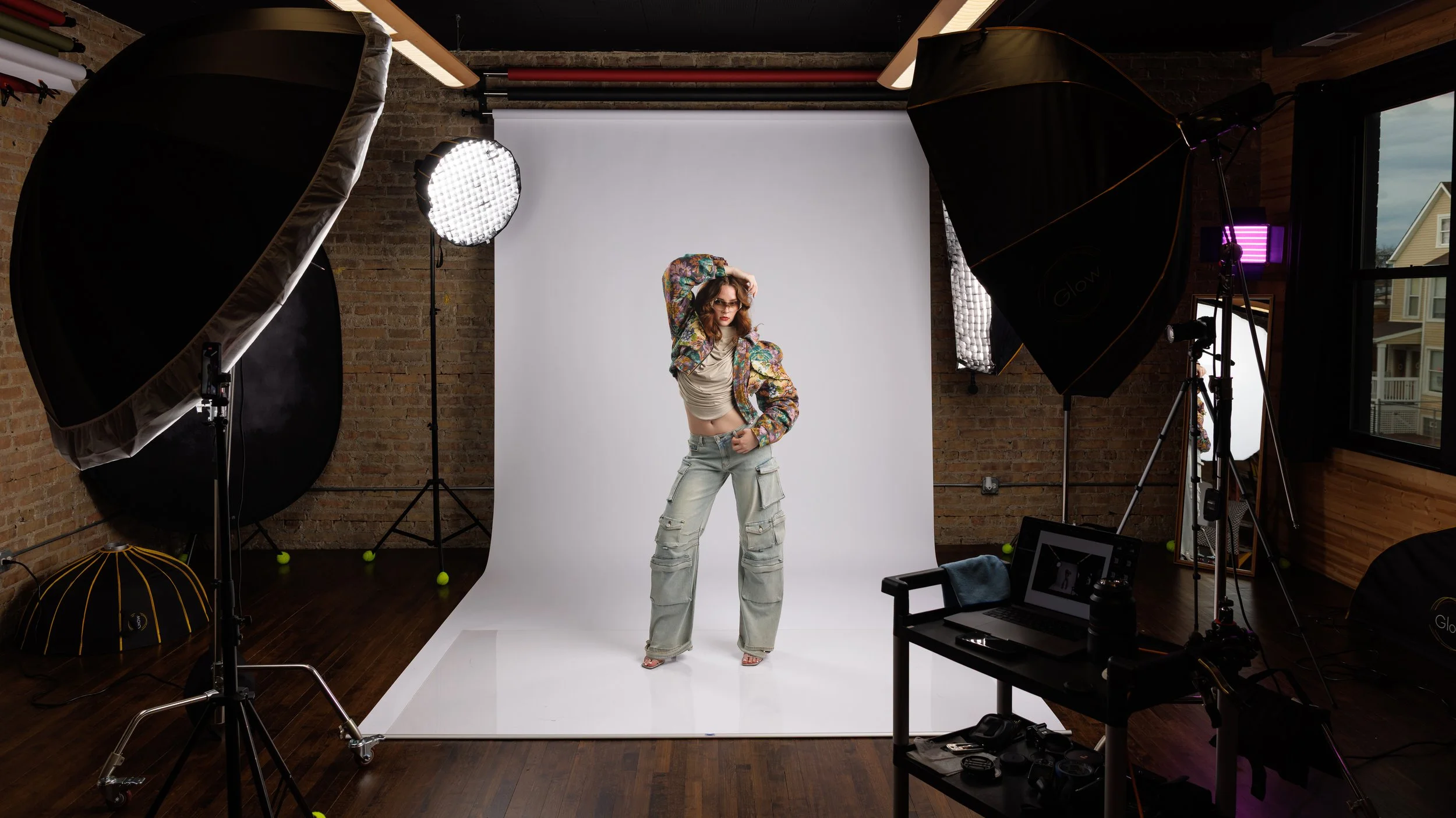 A behind-the-scenes portrait of a studio fashion photoshoot with a model striking a pose in front of a white backdrop, surrounded by professional lighting equipment and camera gear.