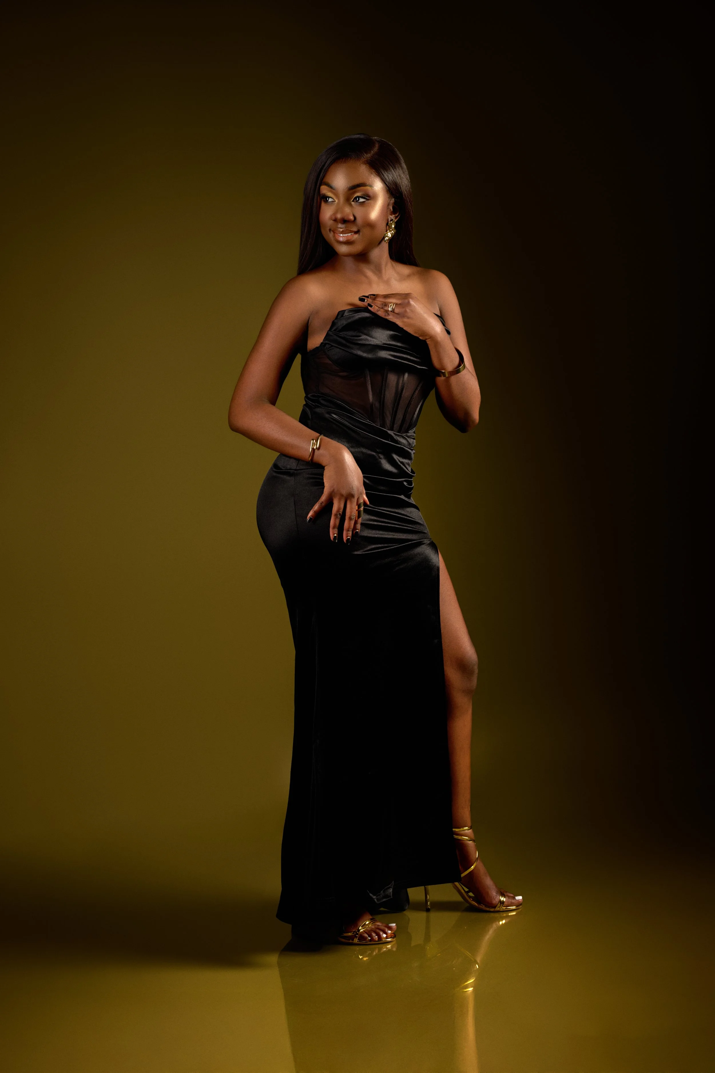 Chicago fashion photoshoot of a female model in a strapless black dress with a high slit, gold high heels, and jewelry, standing against a dark background.