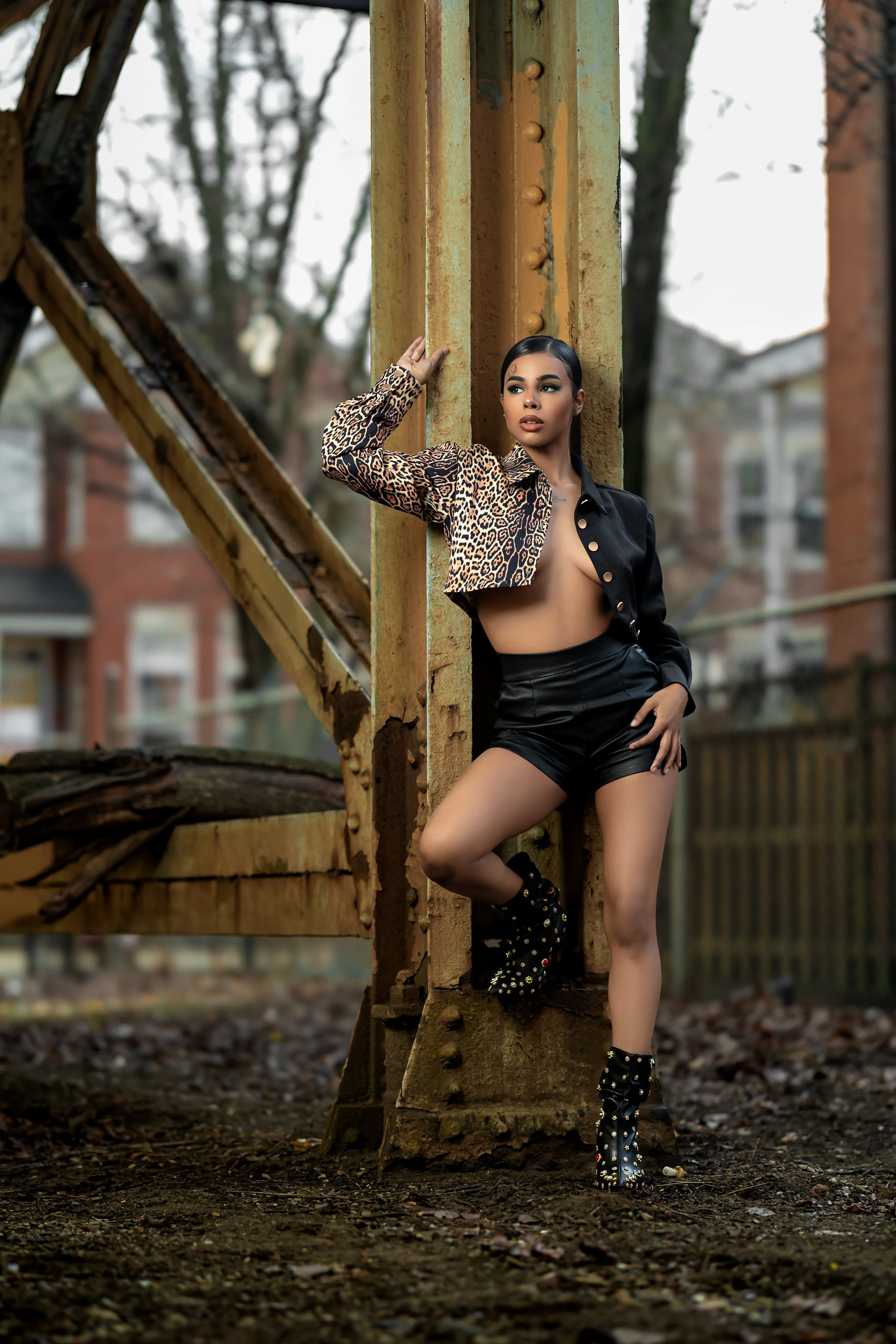A woman in a leopard print jacket, black shorts, and studded heeled boots posing against a rusty metal structure outdoors, with a blurred background of trees and houses.