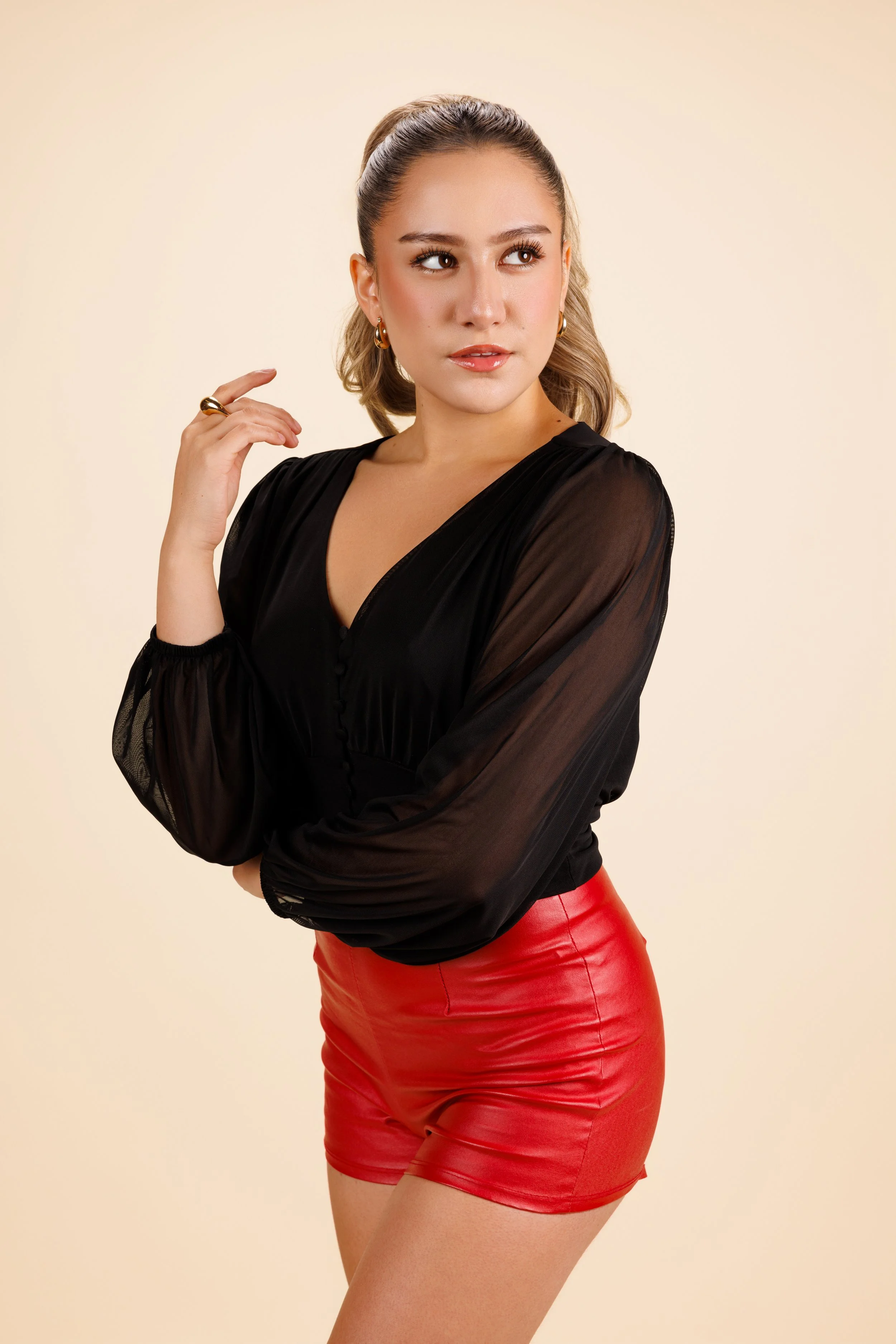A young woman with blonde hair in a ponytail, wearing a sheer black blouse and red leather shorts, posing with one hand near her face against a beige background.