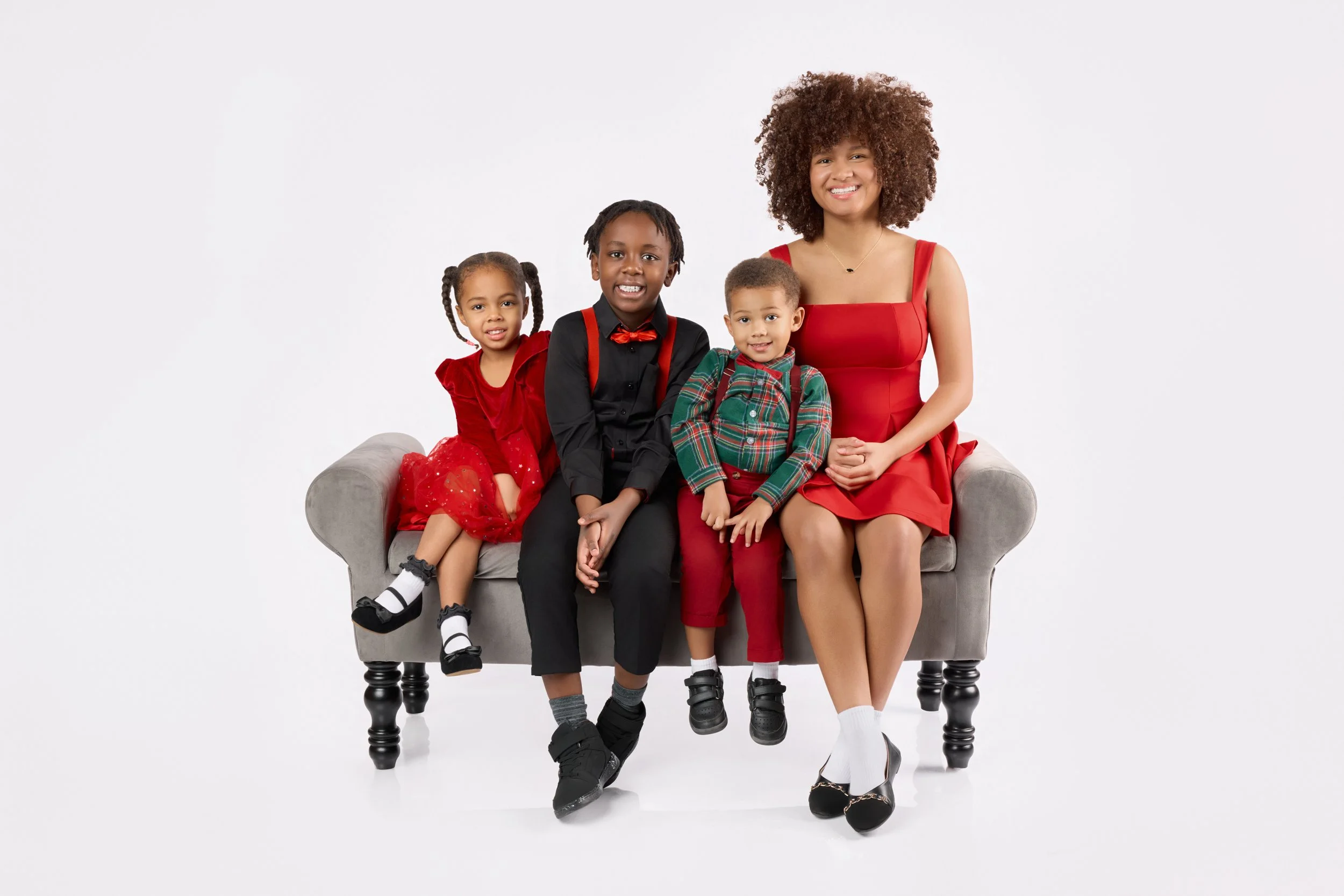 A woman and three children sitting on a gray couch in a studio with white background. The woman and children are dressed in Christmas-themed outfits, and everyone is smiling.